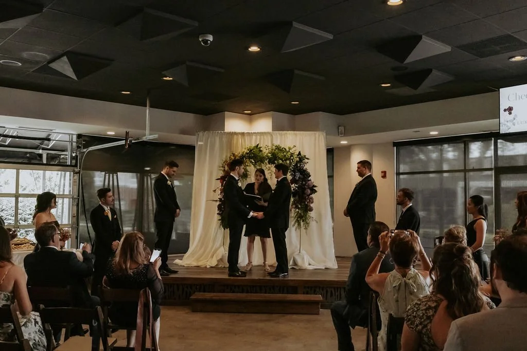 A wedding ceremony taking place indoors with officiants and a bride and groom standing at the altar. Guests seated and standing around the small stage, some using phones. Decor includes flowers and a decorative backdrop.