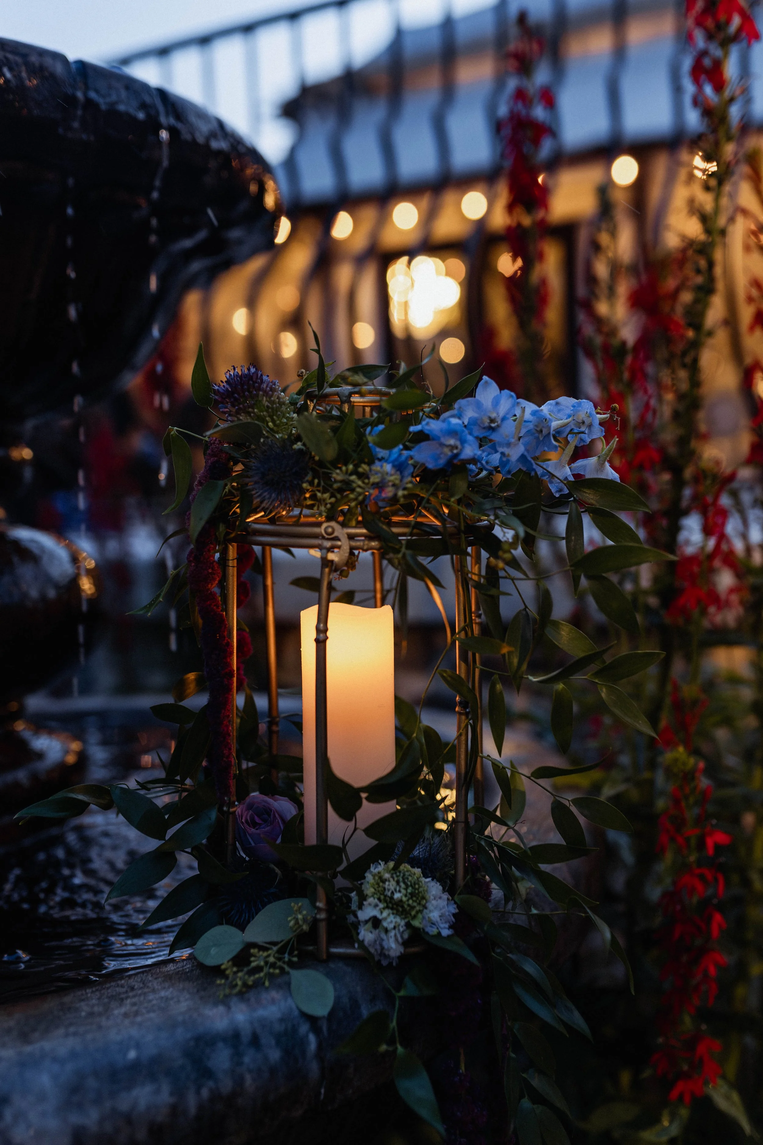 A lit candle surrounded by flowers and greenery at dusk, with a blurred background of string lights and floral decorations.