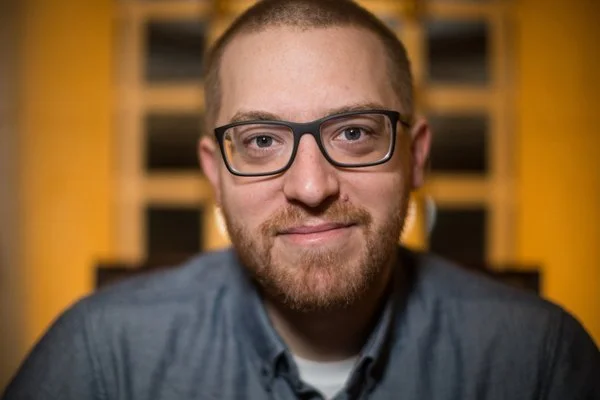 Person in a blue shirt and glasses positioned in front of a yellowish wall.