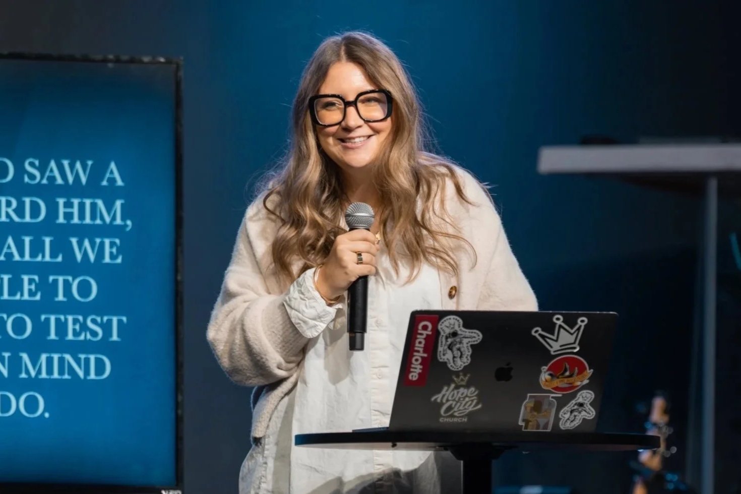 Woman with long wavy hair, glasses, and a beige cardigan, smiling and holding a microphone, standing in front of a laptop with stickers, in a church or presentation setting.