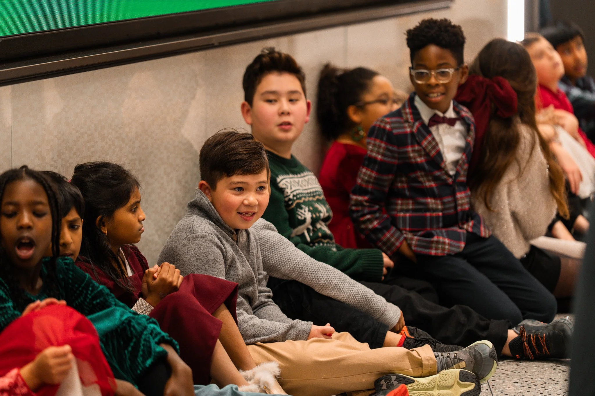 Group of children sitting on the floor against a wall, some wearing festive holiday clothing, smiling and chatting.