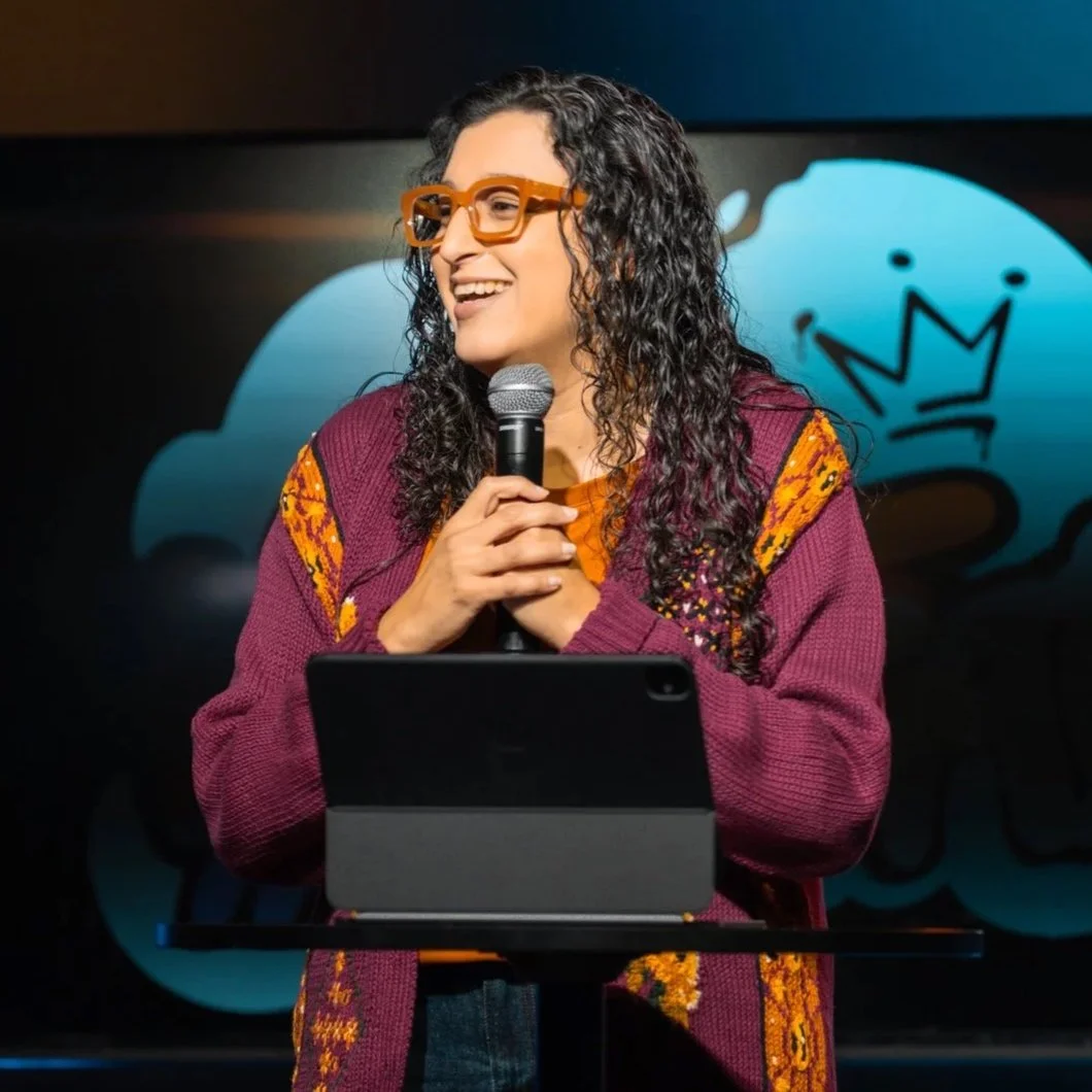 Woman with curly hair and orange glasses speaking into a microphone during a presentation, standing in front of a screen with a blue cloud and black crown graphic.