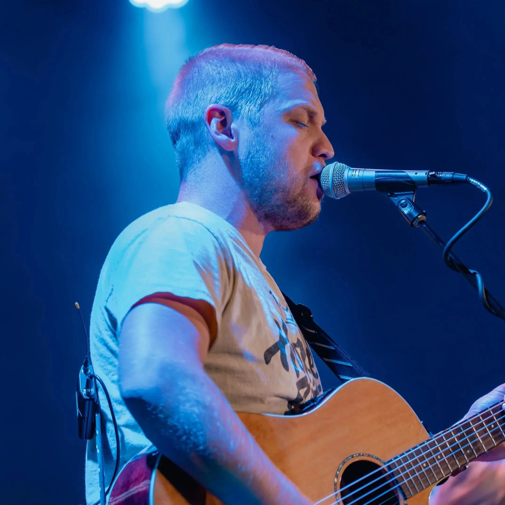 A man in a beige t-shirt playing an acoustic guitar and singing into a microphone on stage, with blue lighting.