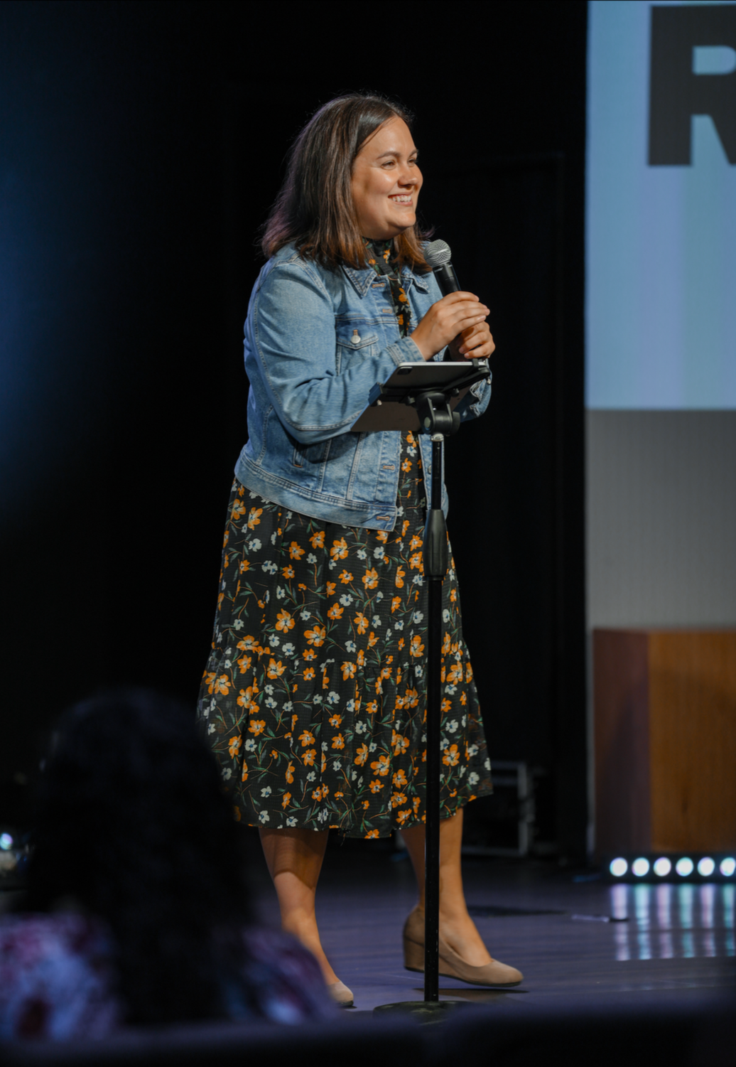 A woman smiling while speaking into a microphone on stage, dressed in a floral dress, denim jacket, and beige heels, with audience in the foreground.