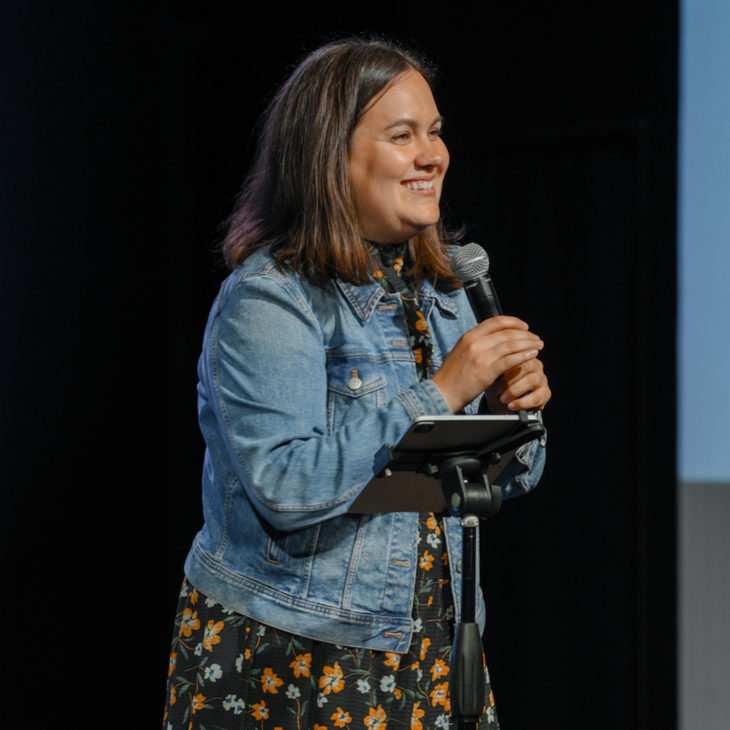 A woman with shoulder-length brown hair, wearing a denim jacket and a black dress with orange and white floral patterns, smiling while holding a microphone and standing in front of a dark background.