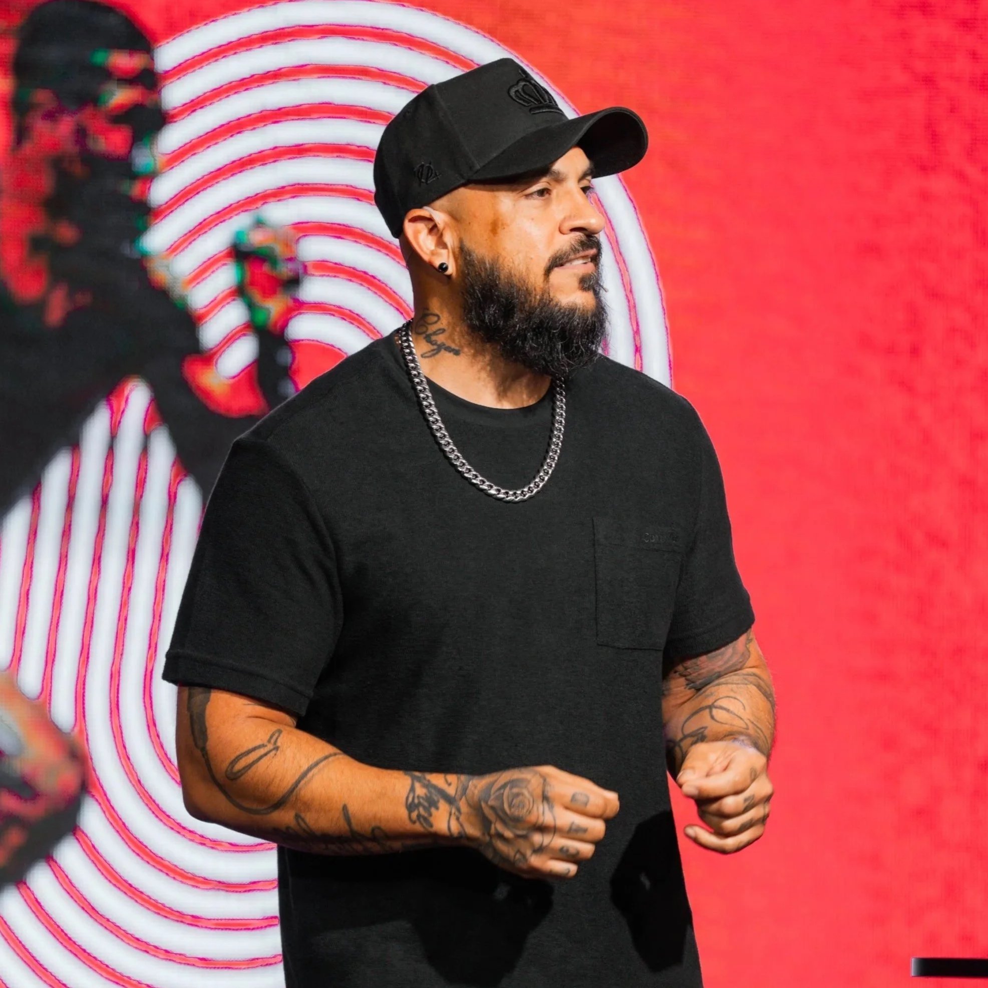 A bearded man wearing a black cap, black T-shirt, silver chain, and tattoos on his arms is standing against a red background with white concentric circles.