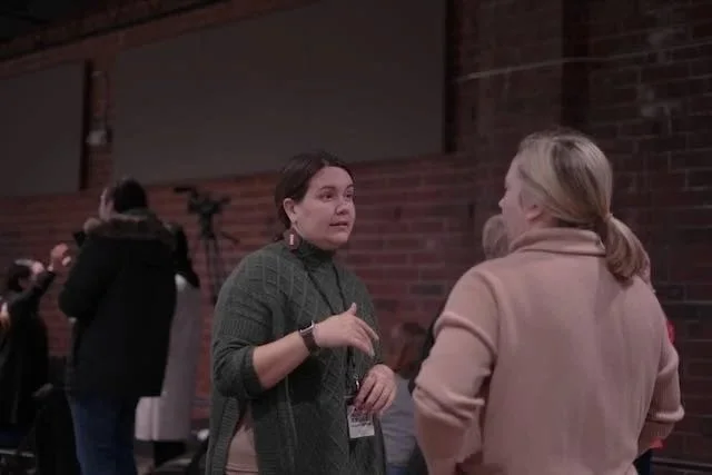Two women are engaged in a conversation at an indoor event with a brick wall in the background. One woman has dark hair, wears a green sweater, and is gesturing with her hand. The other woman has blonde hair, wears a beige coat, and is listening. Several people are visible in the background.