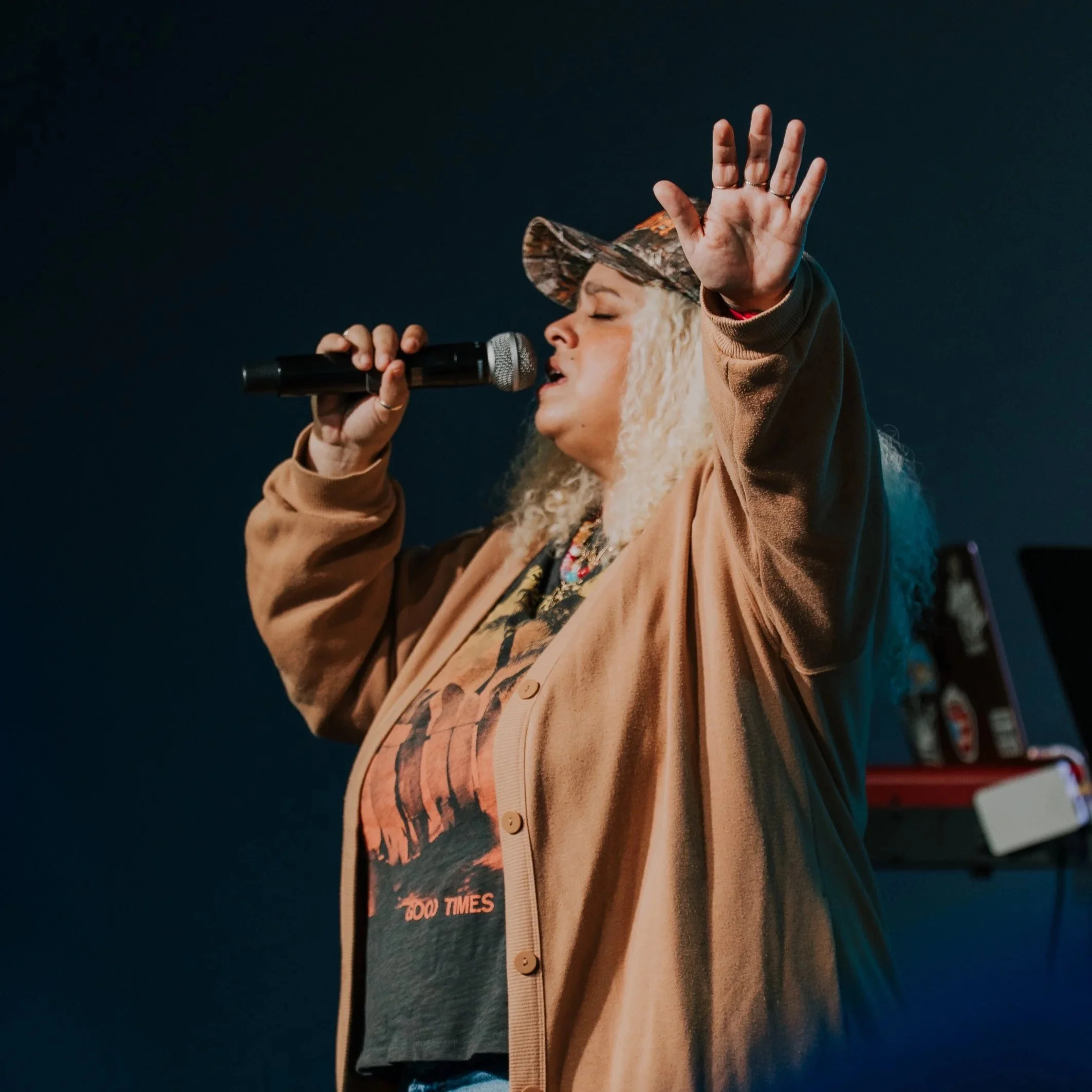 A woman with blonde, curly hair singing into a microphone with her eyes closed, wearing a beige jacket and a camo cap, on stage with a dark background.