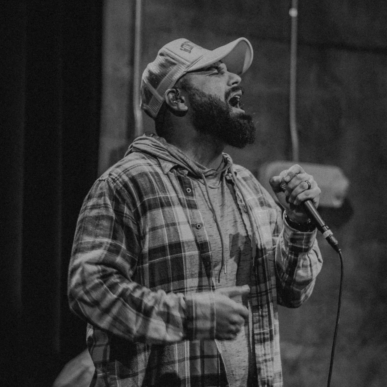 A man with a beard singing passionately into a microphone while wearing a baseball cap, hoodie, and plaid shirt in a dark setting.