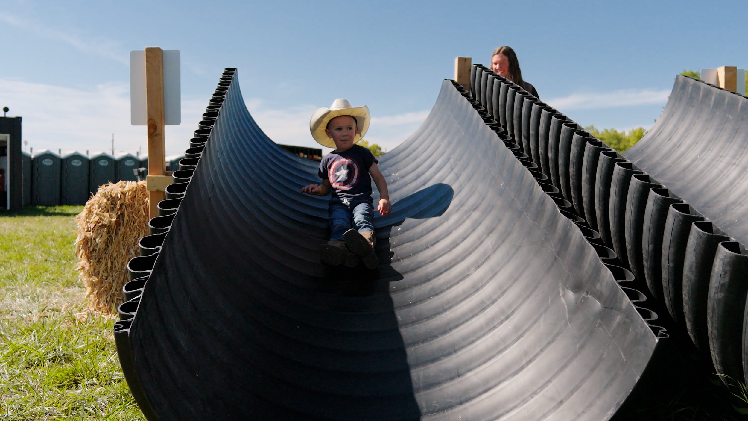 A young boy wearing a cowboy hat and a woman are riding down a large, black, curved pipe slide at an outdoor fair or festival, with grass in the background.
