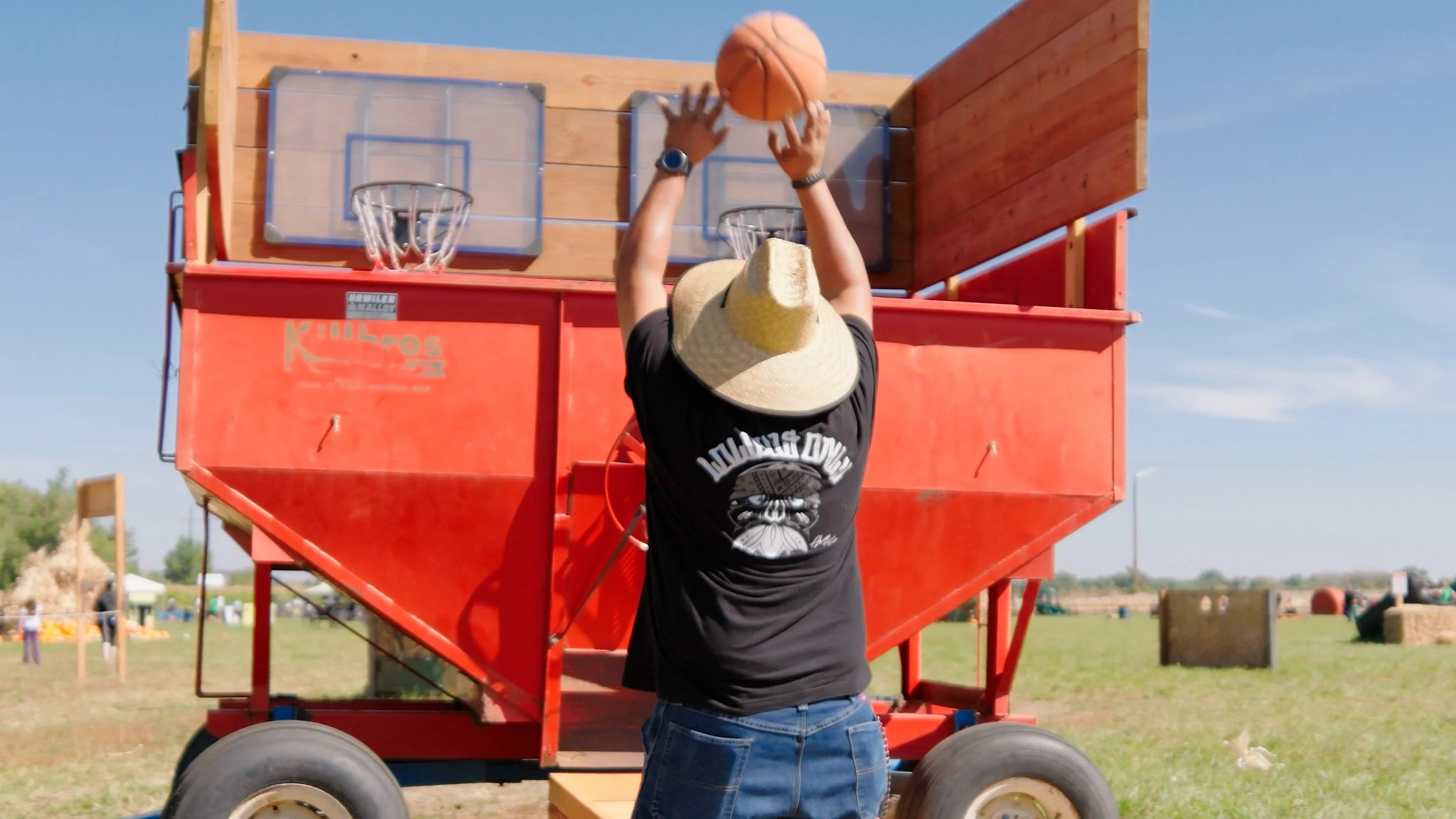 A person wearing a hat and dark t-shirt playing basketball on an outdoor court with a red wooden basketball shooting game in the background.
