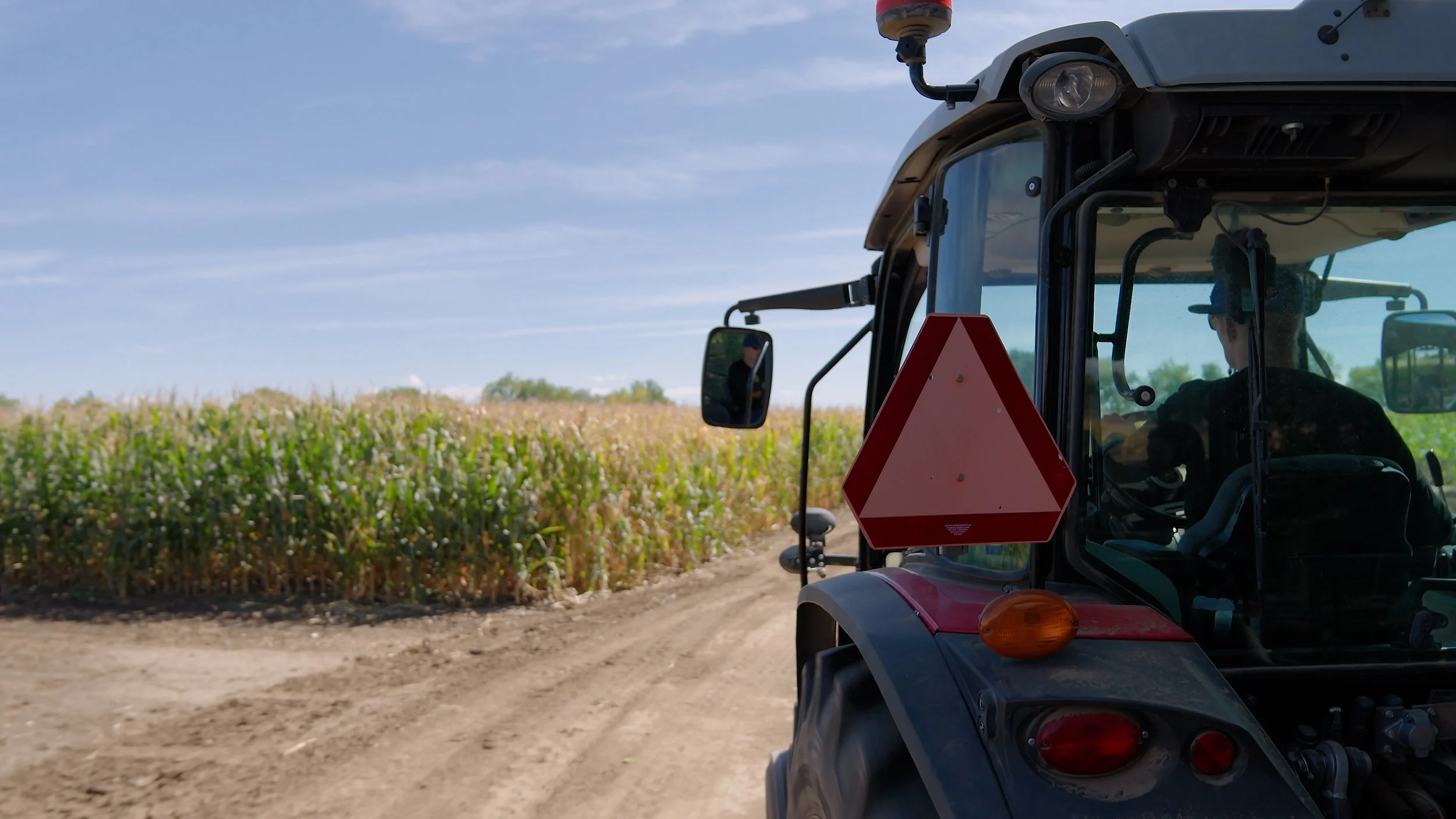 A tractor driving along a dirt farm road in front of a large field of plants, with a blue sky and some clouds in the background.