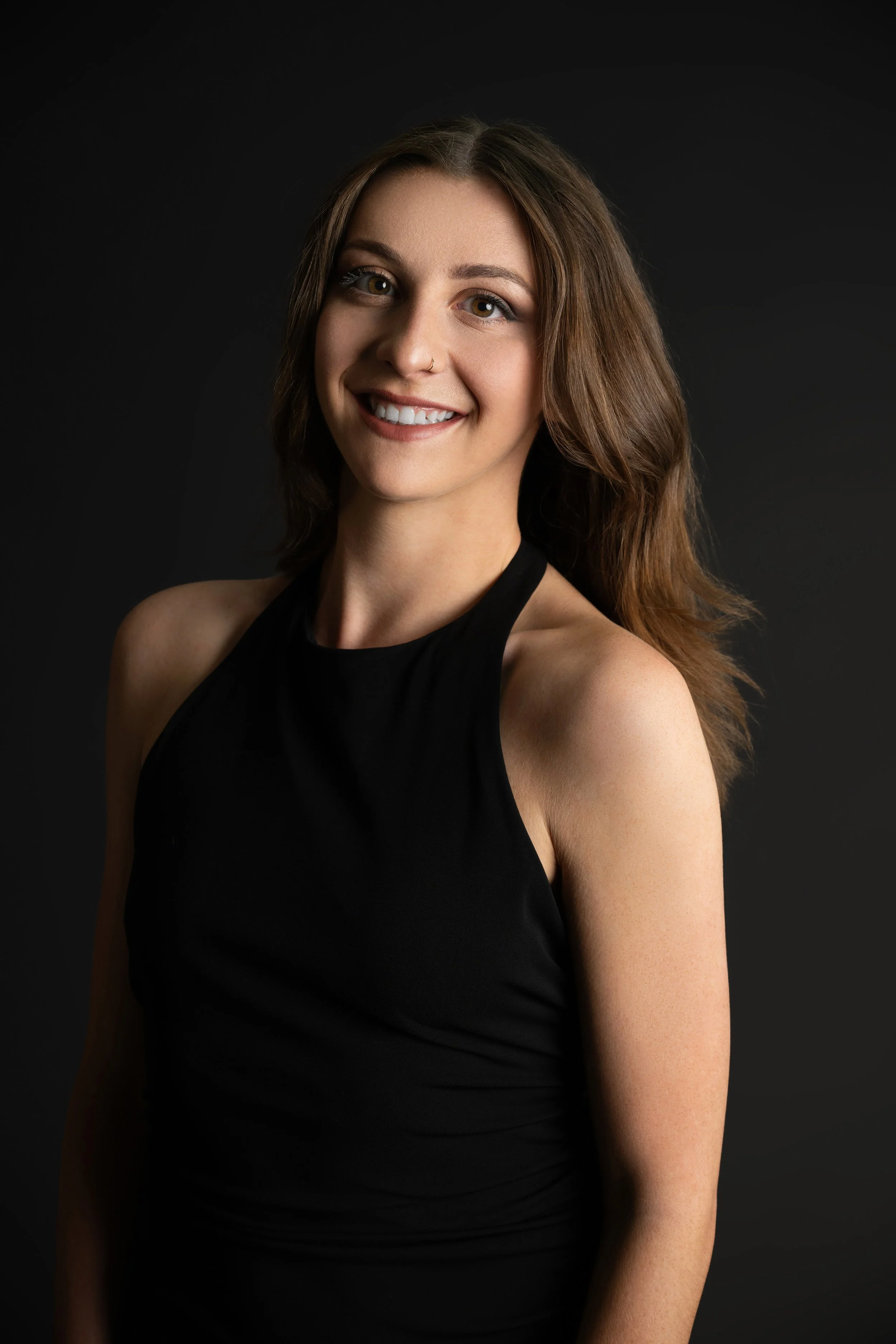 Portrait of a young woman with long brown hair, smiling, wearing a black sleeveless top, against a dark background.