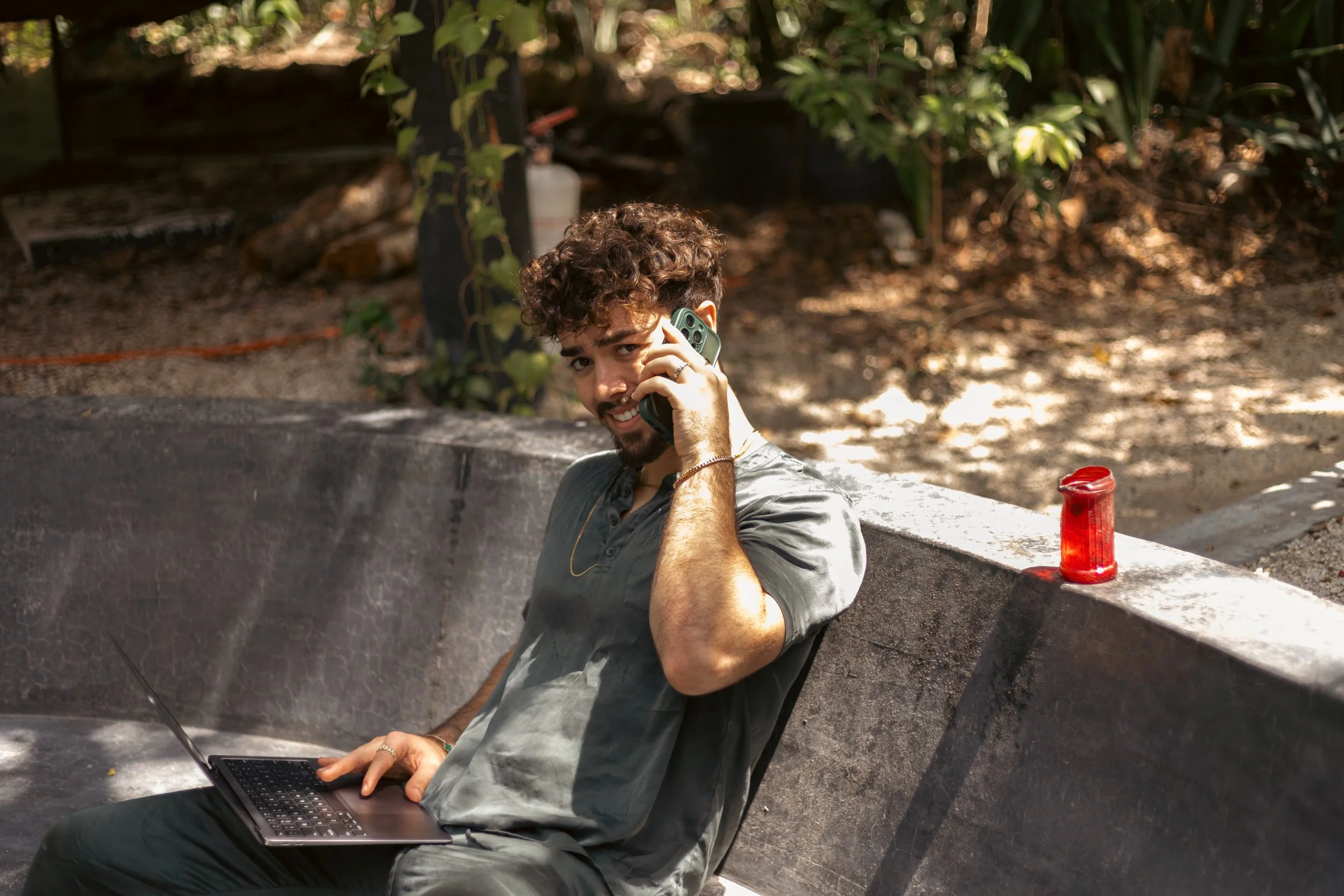 Man sitting outdoors on a curved bench, holding a smartphone to his ear and using a laptop on his lap, with a red drink container on the bench beside him.