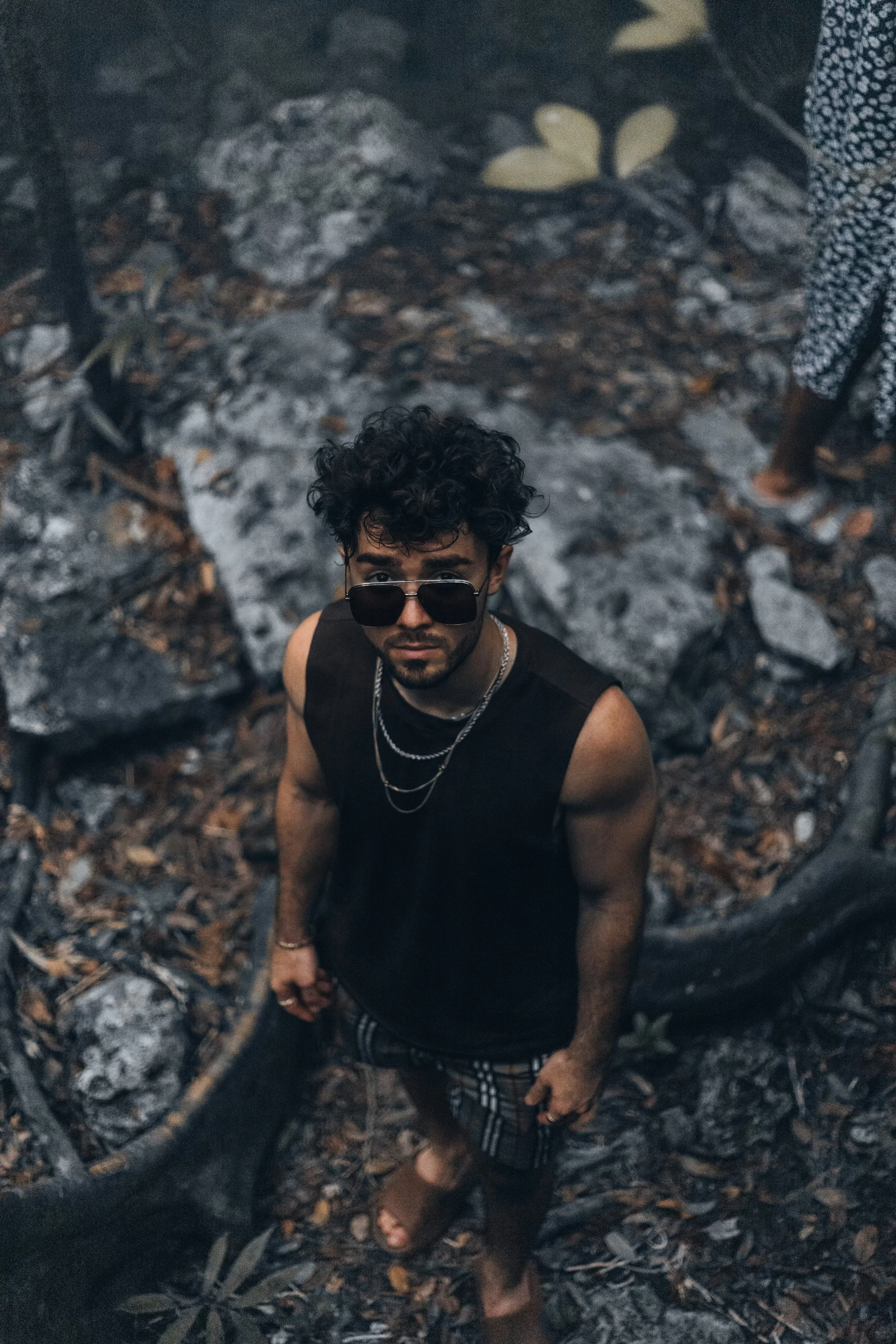 Man wearing sunglasses and a sleeveless shirt stands outdoors on rocky terrain looking up.