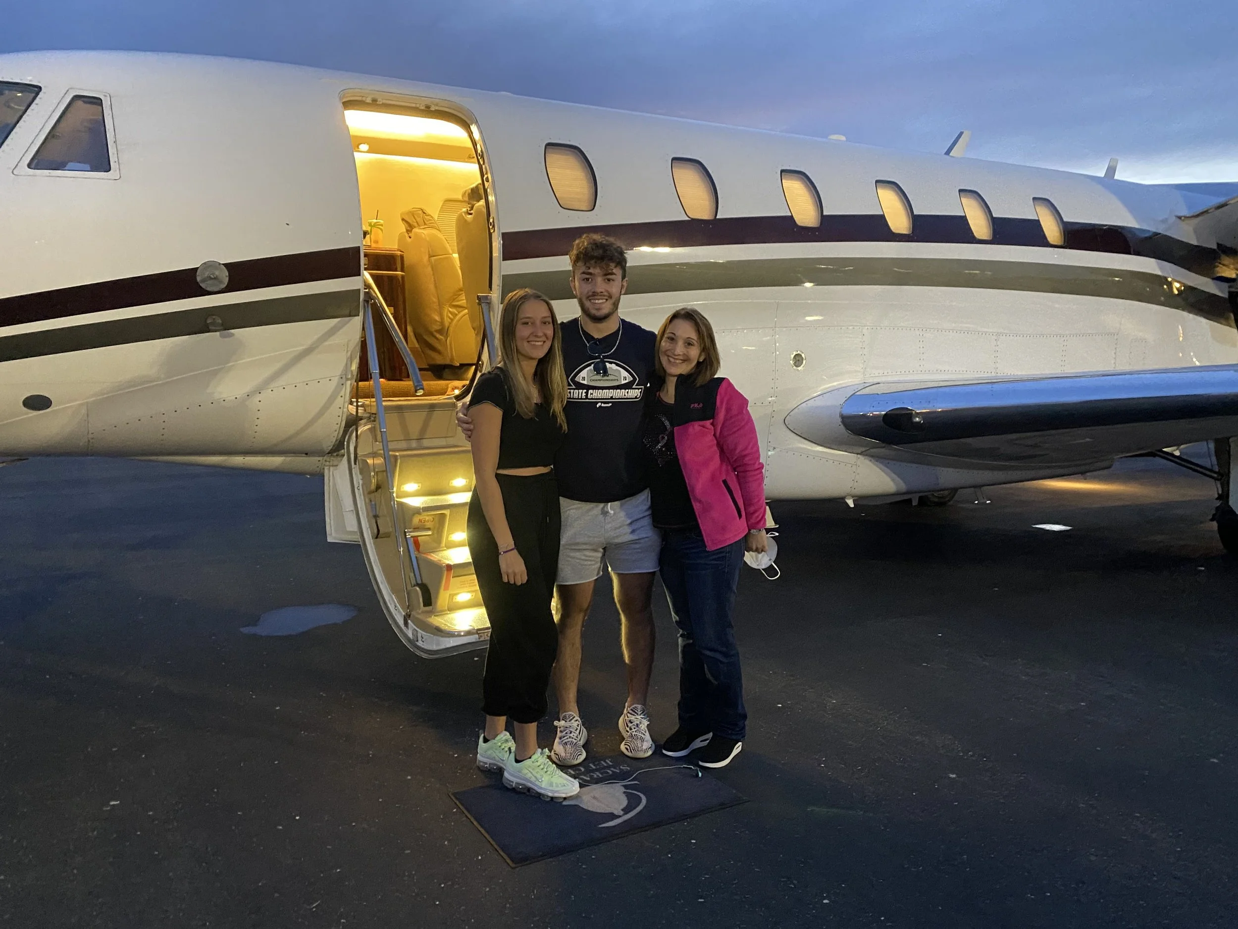 Three people standing in front of a private jet on an airport tarmac in the evening.