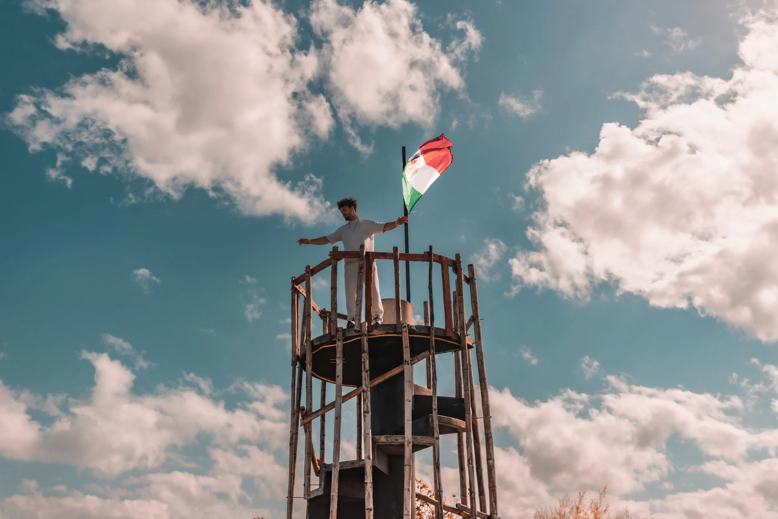 Person standing on top of a wooden tower holding a Mexican flag against a blue sky with clouds.