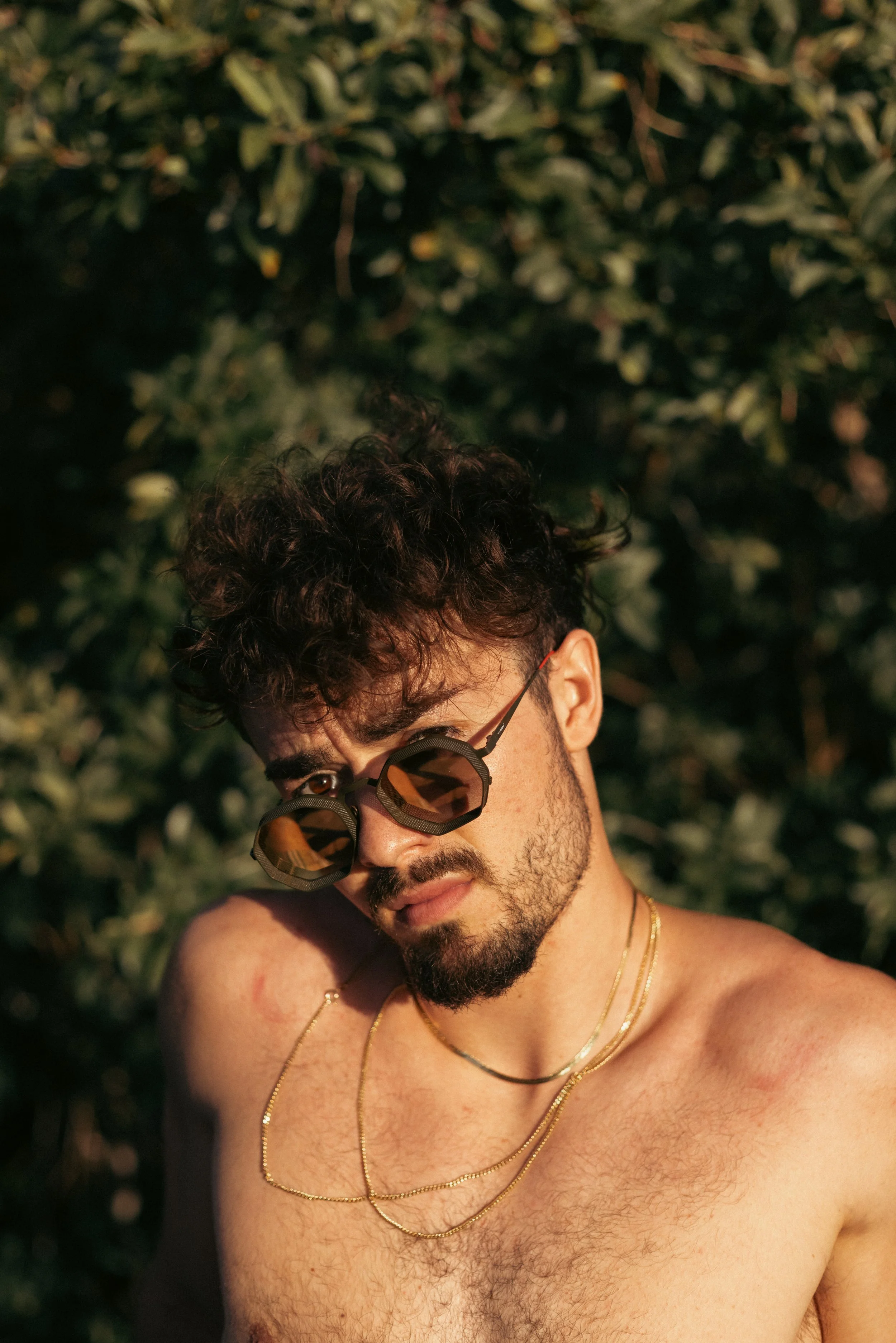 Man with curly hair and sunglasses posing outdoors in front of leafy background, wearing gold necklaces, shirtless.