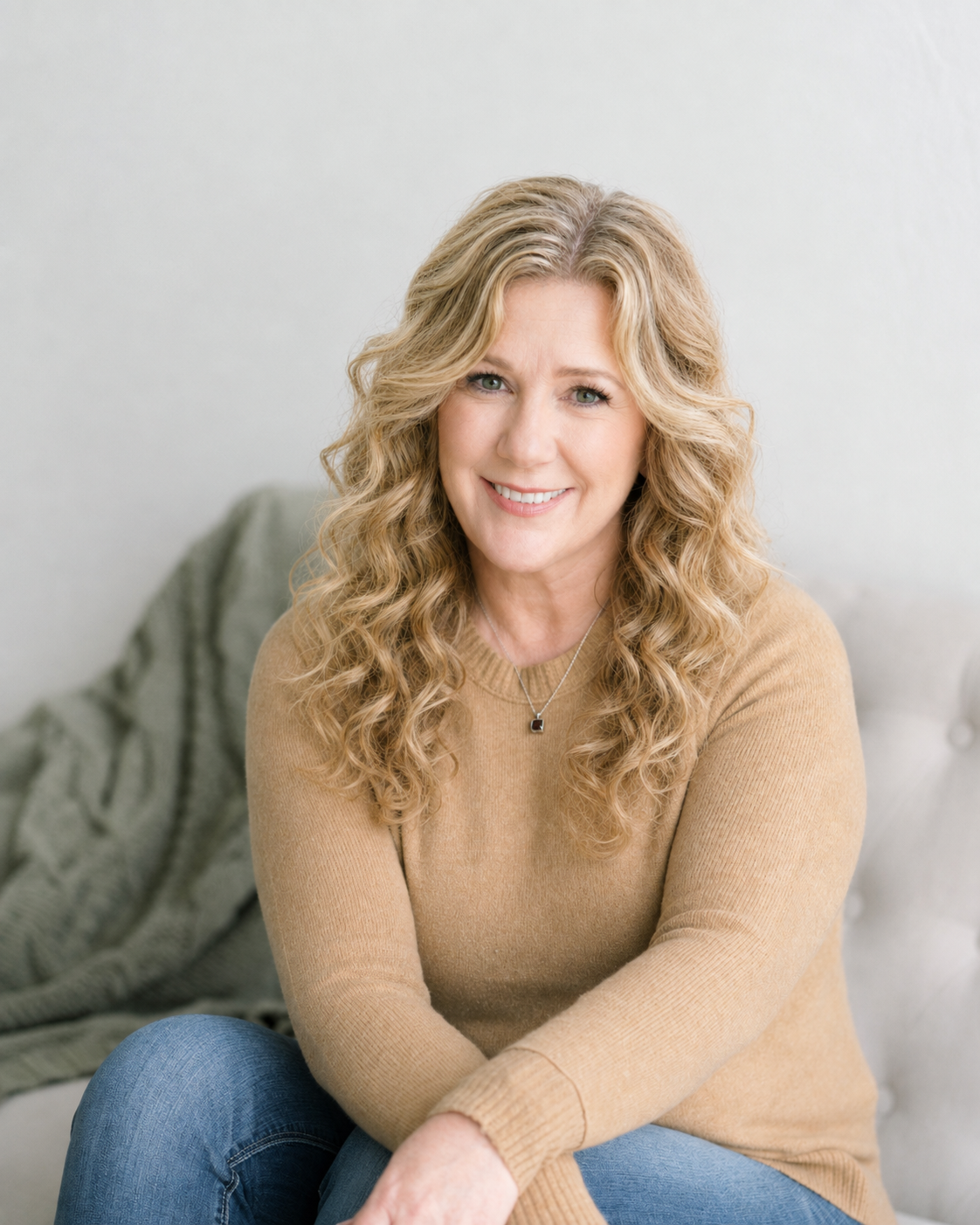 Michelle Lanlgey, LPC smiling at the camera, wearing a tan sweater and blue jeans, sitting indoors with a light-colored wall and a green blanket in the background.