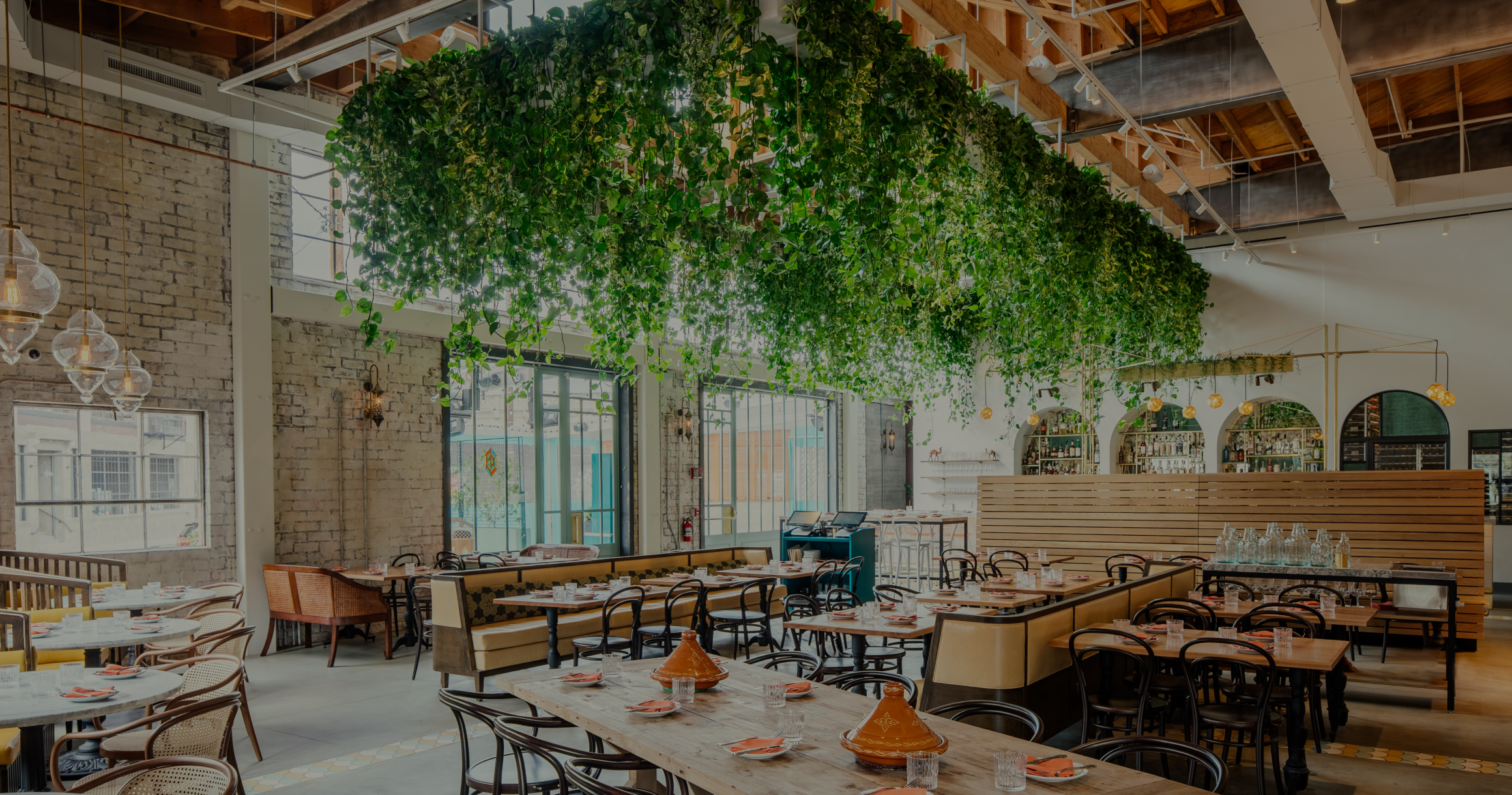 Interior of a modern restaurant with hanging plants, wooden tables, chairs, and pendant lights, featuring empty plates and glasses on the tables.