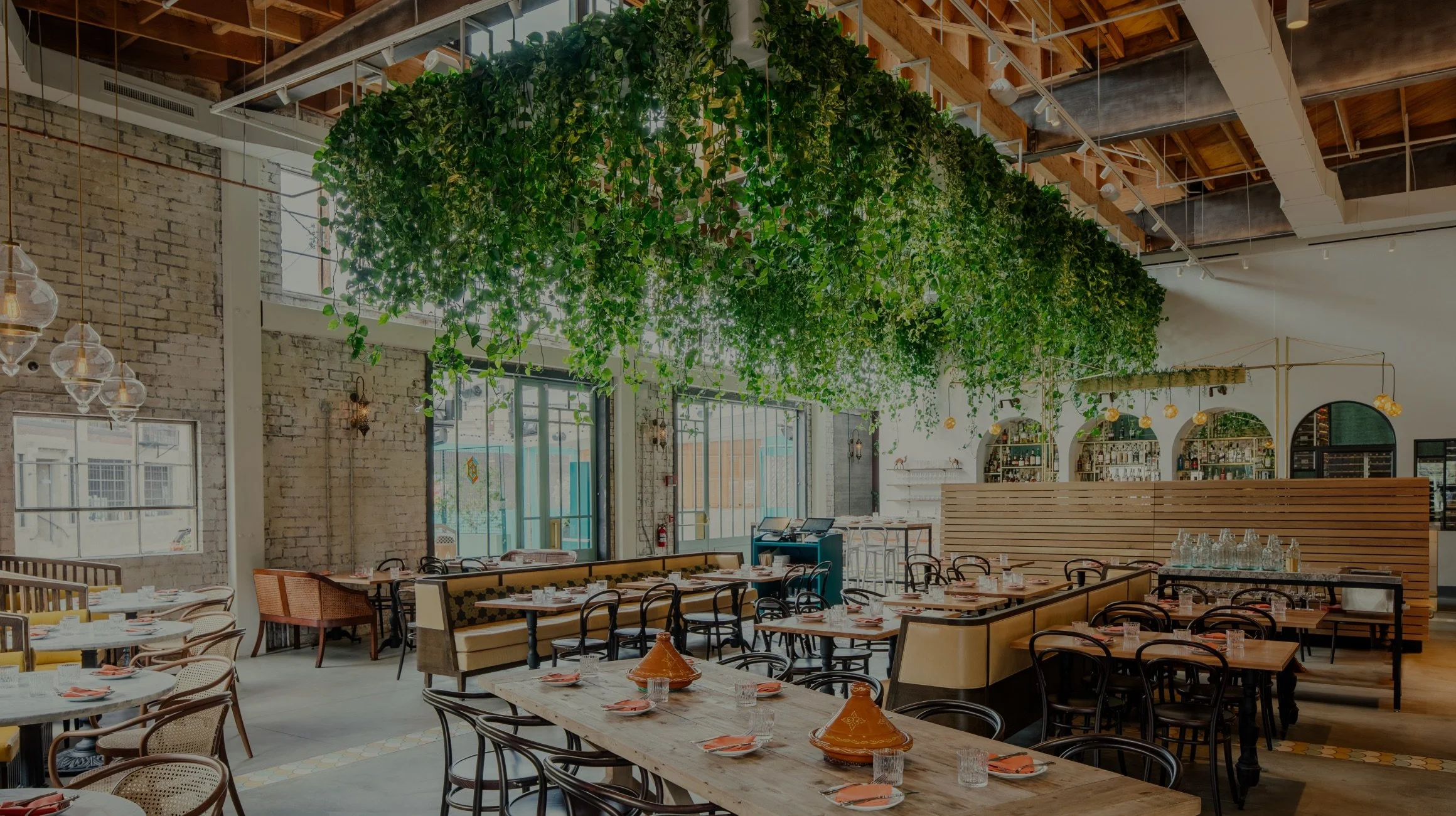 Stylish restaurant interior with wooden tables, chairs, and suspended greenery. Exposed brick walls and modern lighting fixtures enhance the contemporary vibe.