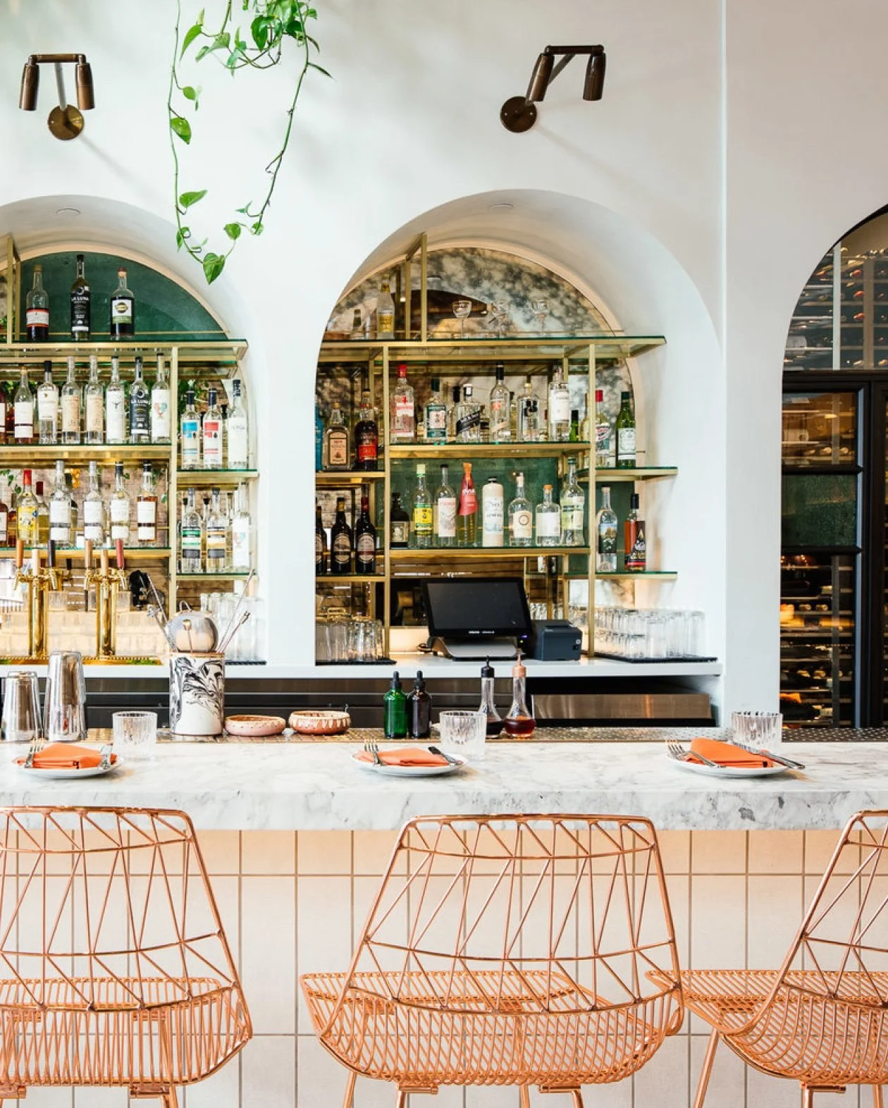 Stylish bar interior with marble countertop, wire metal chairs, and arched shelves displaying various bottles of alcohol. Minimalist decor includes potted plant and dining setup with plates and glasses.