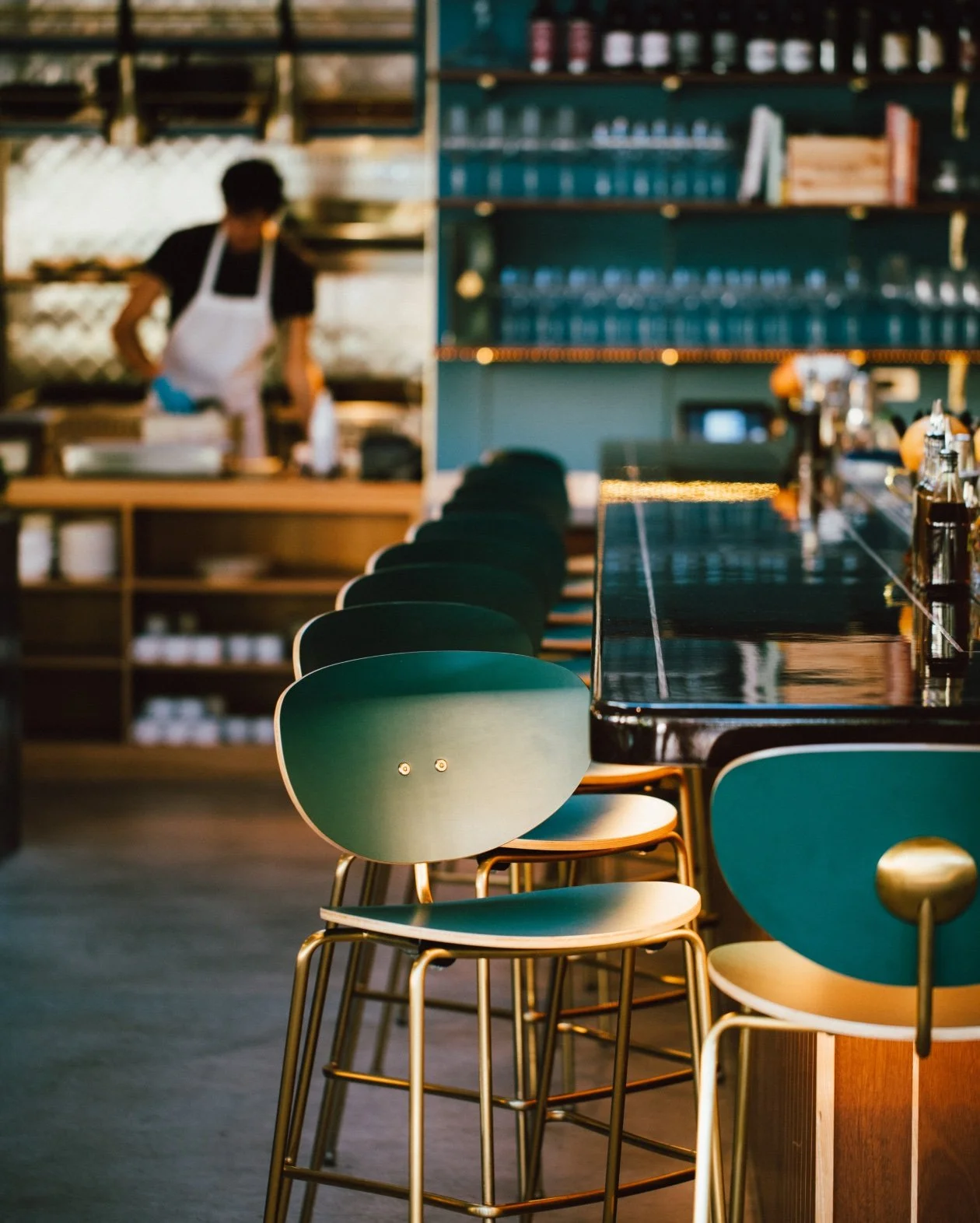 Interior of a modern bar with green bar stools lined up at a glossy dark bar counter, shelves with glassware and bottles in the background, and a person in an apron working in the kitchen area.