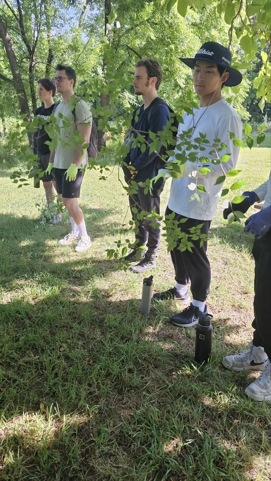 Group of young people standing outdoors in a park, some wearing gloves and holding water bottles, gathered under the shade of trees.