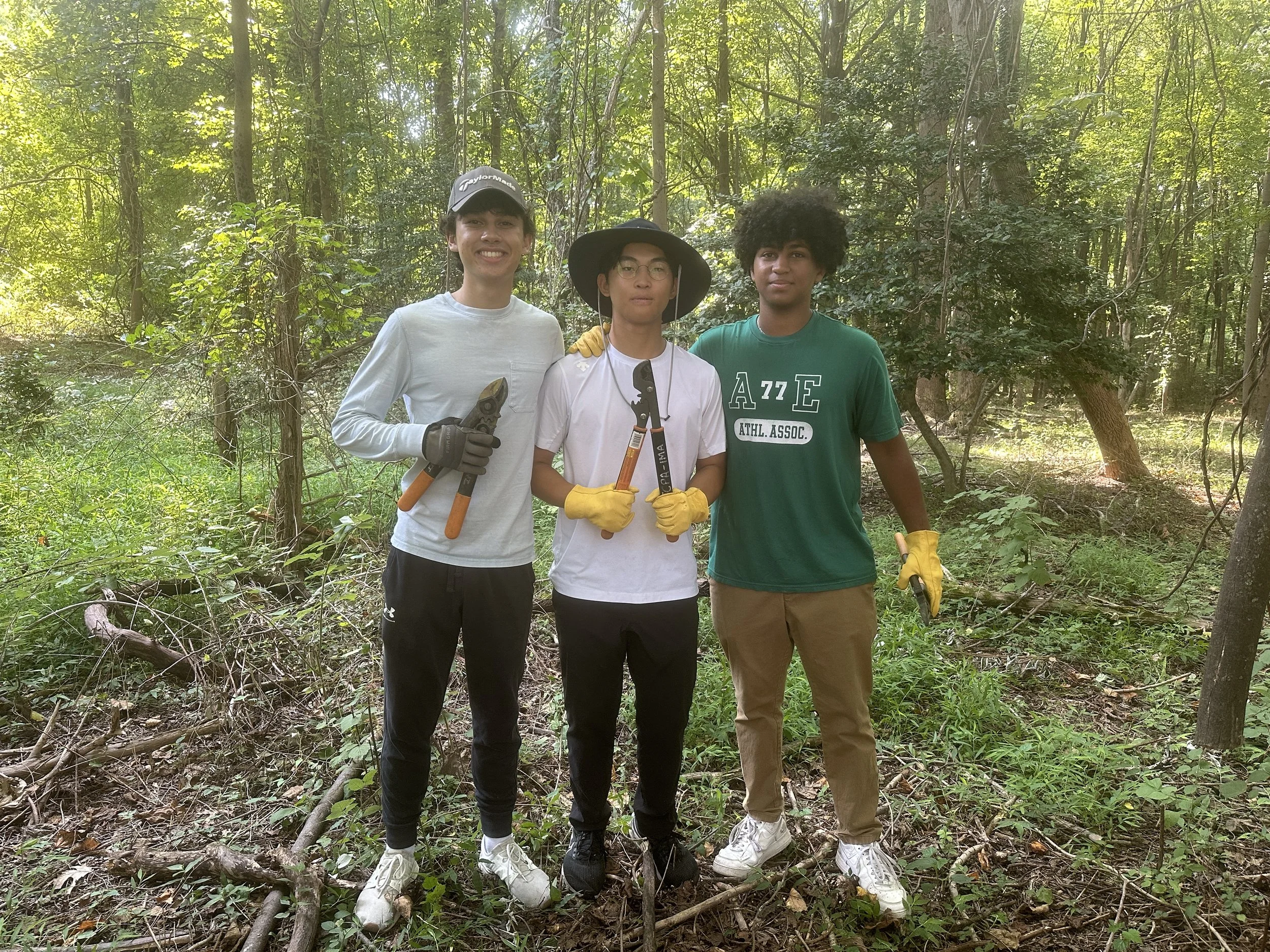 Three young men standing together in a lush forest. They are dressed casually, with gloves and holding pruning or gardening tools, indicating they may be working on a conservation or planting activity.
