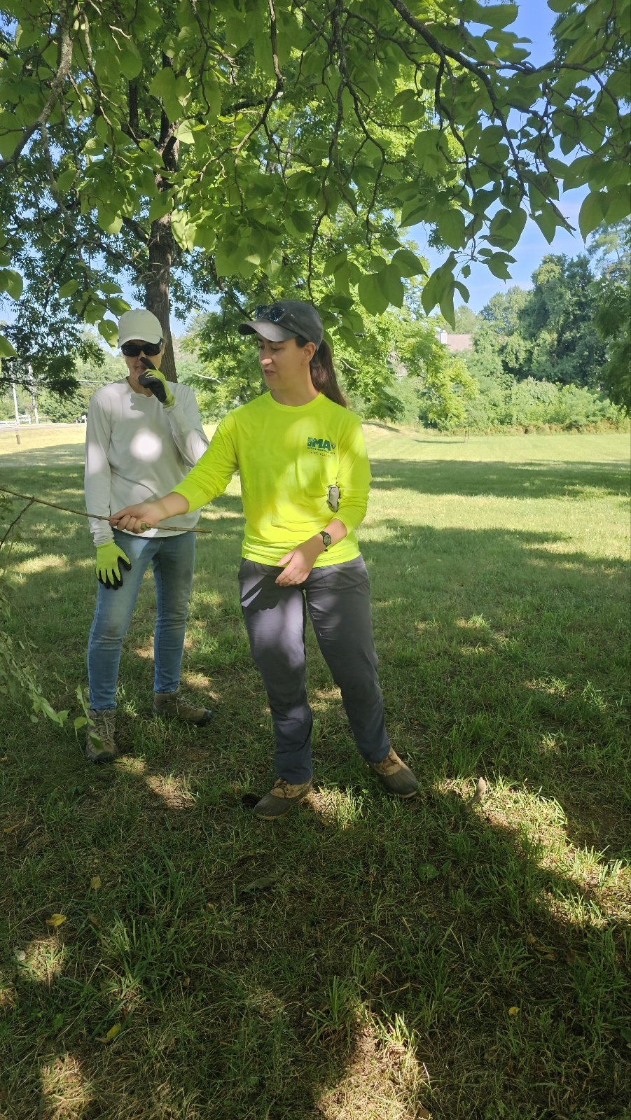 Two women standing under a tree in a grassy park. One woman in a bright yellow shirt appears to be explaining something, while the other in a white shirt and cap listens, both wearing gloves.
