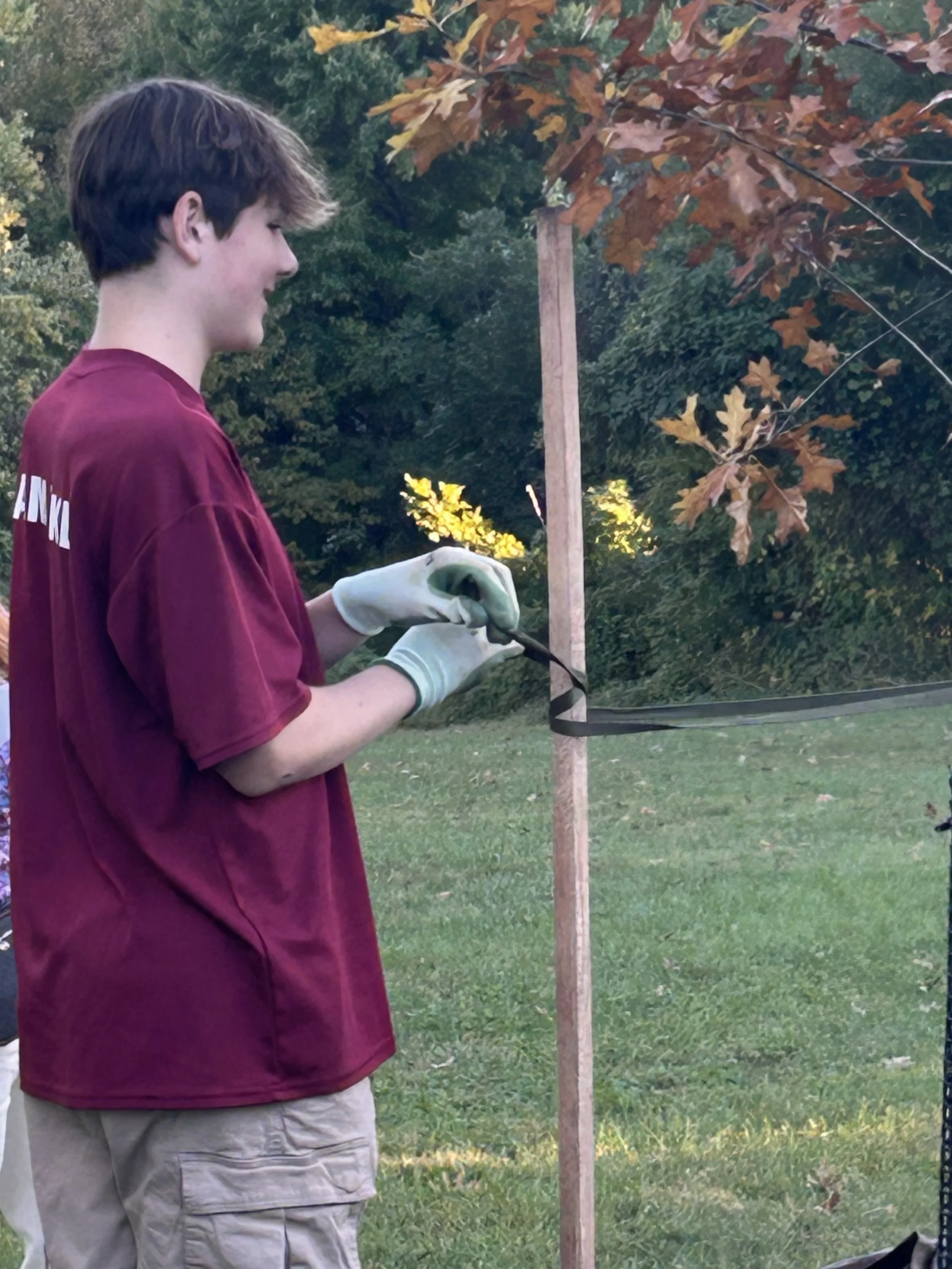A young man wearing a maroon T-shirt and beige cargo shorts is using gloves and a tool to cut a strip of landscape fabric around a wooden stake in a grassy yard, with trees and bushes in the background.