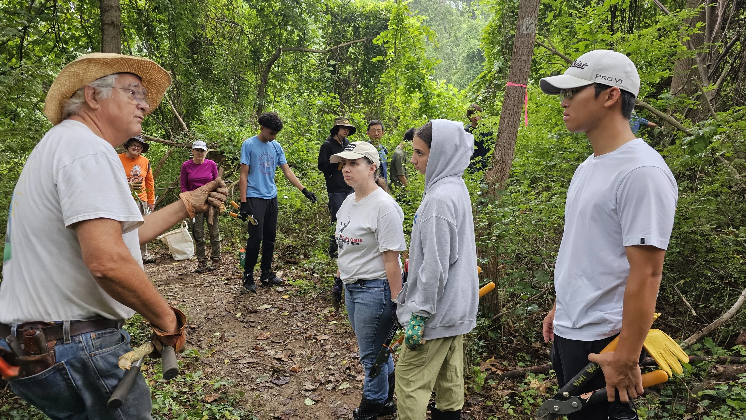 Group of people in a forest, listening to an older man with a straw hat, who appears to be giving a demonstration or lecture about nature or gardening. Some individuals are holding tools, and the setting is lush with green foliage.