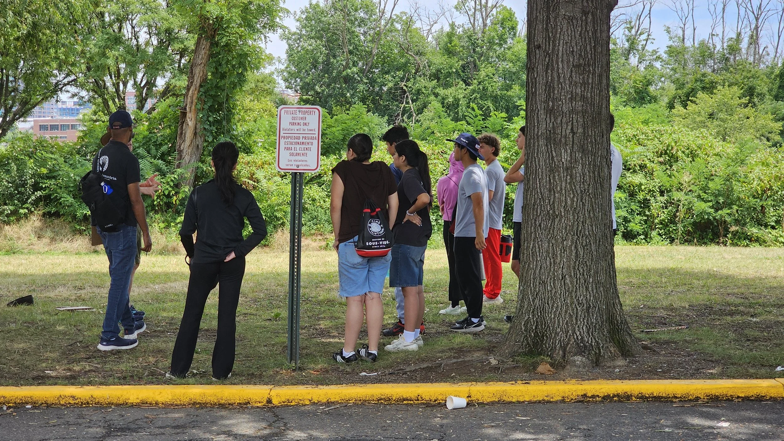 Group of people standing outdoors near trees, listening to a man speak, with a red and white sign that says 'Private Property' in the background.