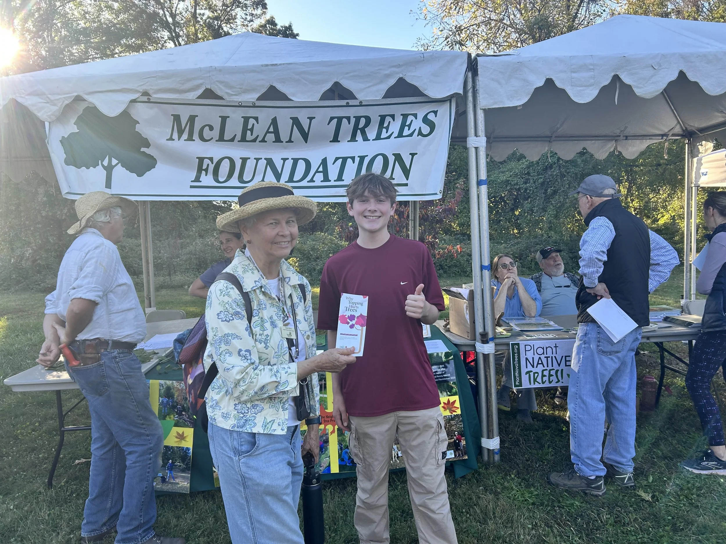 A woman in a floral shirt and straw hat holding a pamphlet, giving a thumbs-up, standing next to a young man in a maroon shirt at an outdoor plant and tree organization booth called McLean Trees Foundation. Several people are seated and standing around the booth under a white canopy with a banner, with trees and grass in the background.