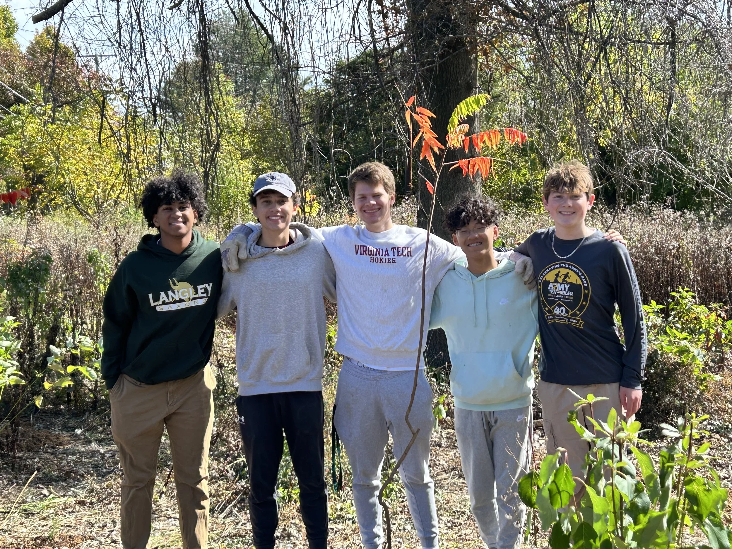 Five young males standing outdoors in a natural setting, smiling with arms around each other's shoulders, with trees and bushes in the background.