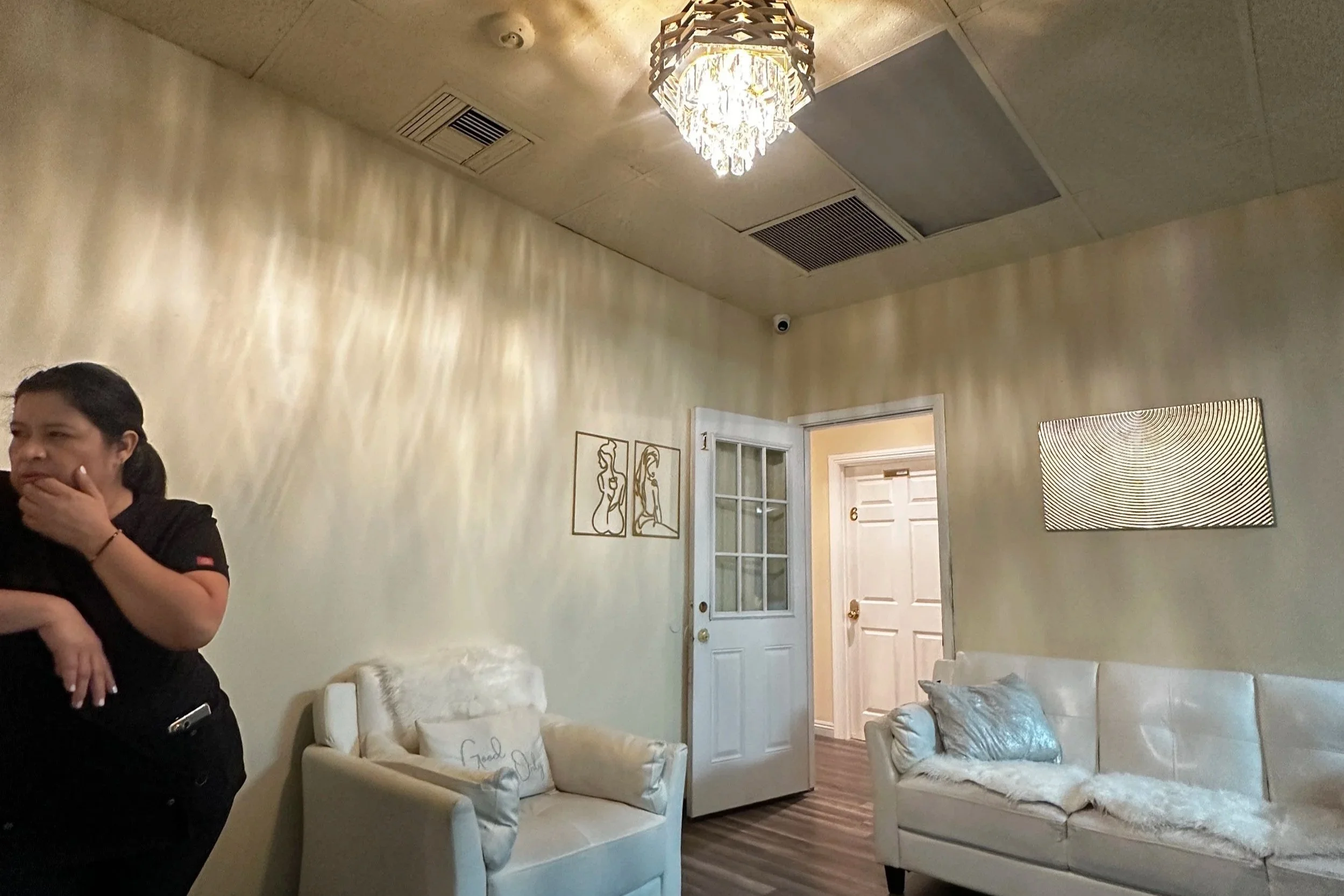 Living room with white sofa, decorative pillows, and a woman standing on the left side. The room has beige walls, a modern chandelier on the ceiling, and a door with a glass window leading to other rooms. Wall art includes a minimalist line drawing of a woman and a textured rectangular mirror.