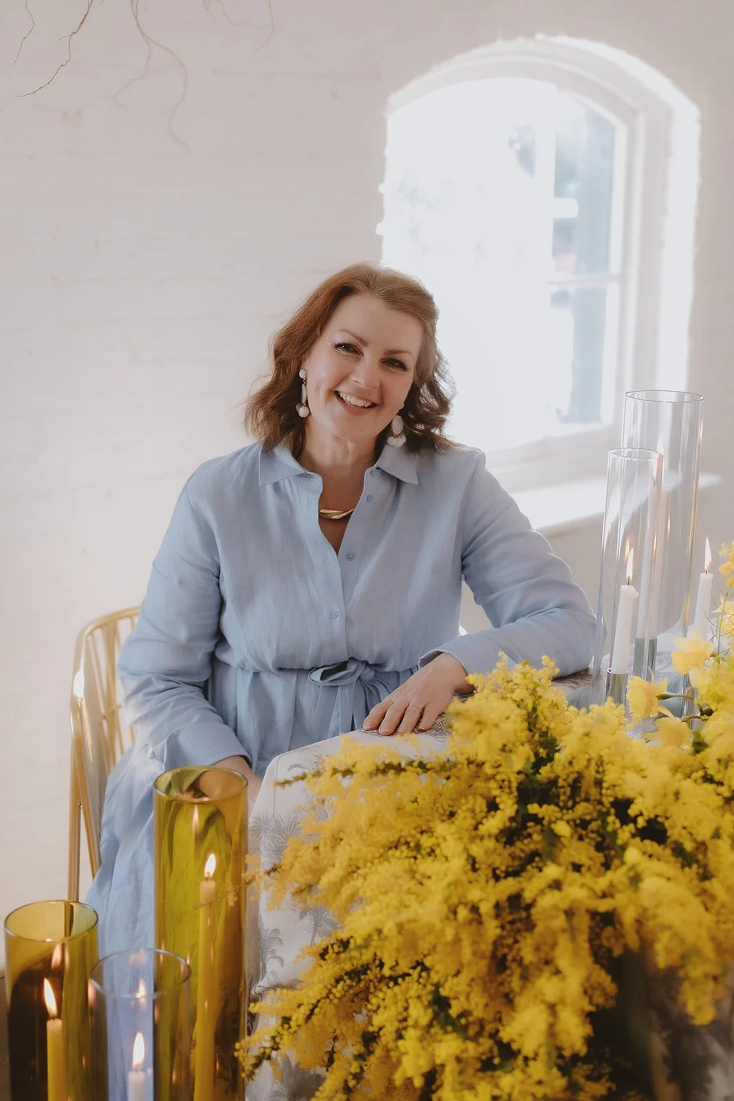 Woman sitting at a table with yellow flowers and lit candles, wearing a light blue dress in a bright room with a window in the background.