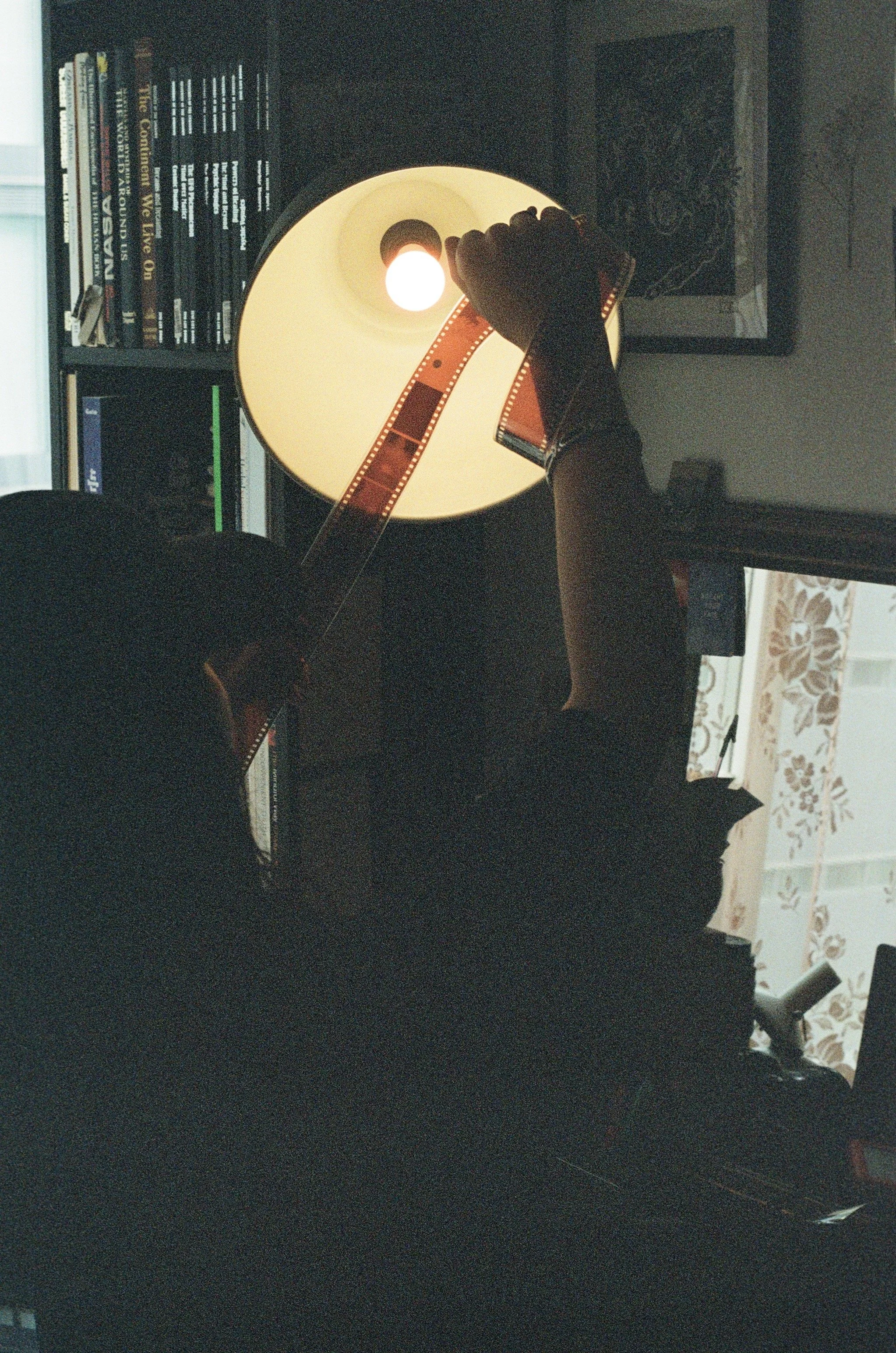 Person holding a strip of film strip in front of a lit lamp on a nightstand, with a bookshelf filled with books in the background.