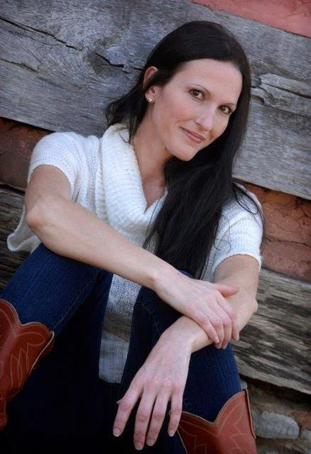 A woman with long dark hair, wearing a white sweater and jeans, sits against a wooden background, posing for a casual outdoor portrait. She is also wearing boots.