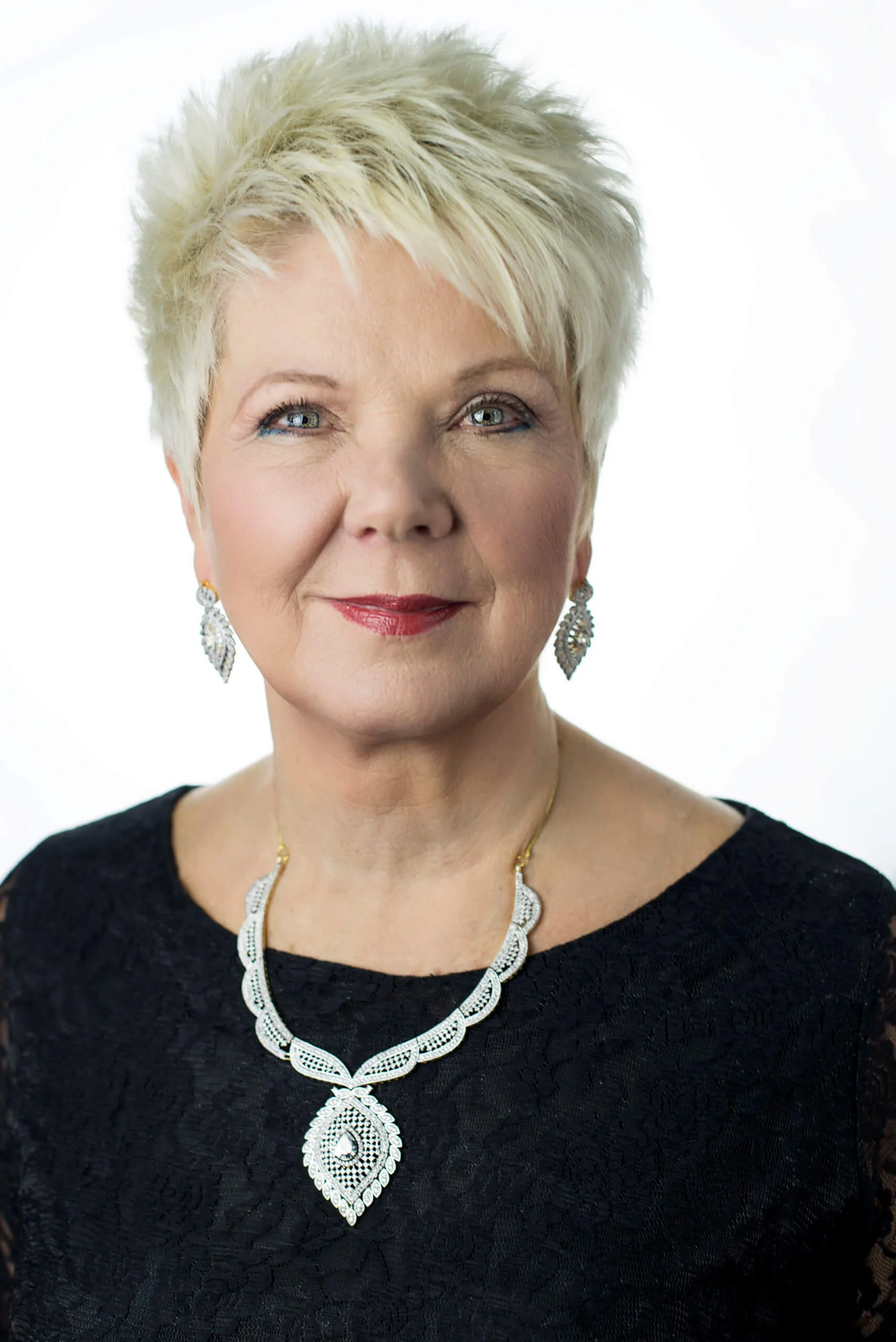 Woman with short blonde hair wearing a black lace top, decorative necklace, and matching earrings, posing against a white background.