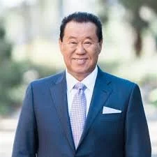 A man in a suit with a light blue tie, smiling outdoors against a blurred background of greenery.