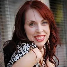 Smiling woman with red hair wearing a patterned top, leaning against a surface with horizontal blinds in the background.