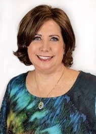 Smiling woman with short brown hair, wearing a colorful top and a necklace, on a white background.