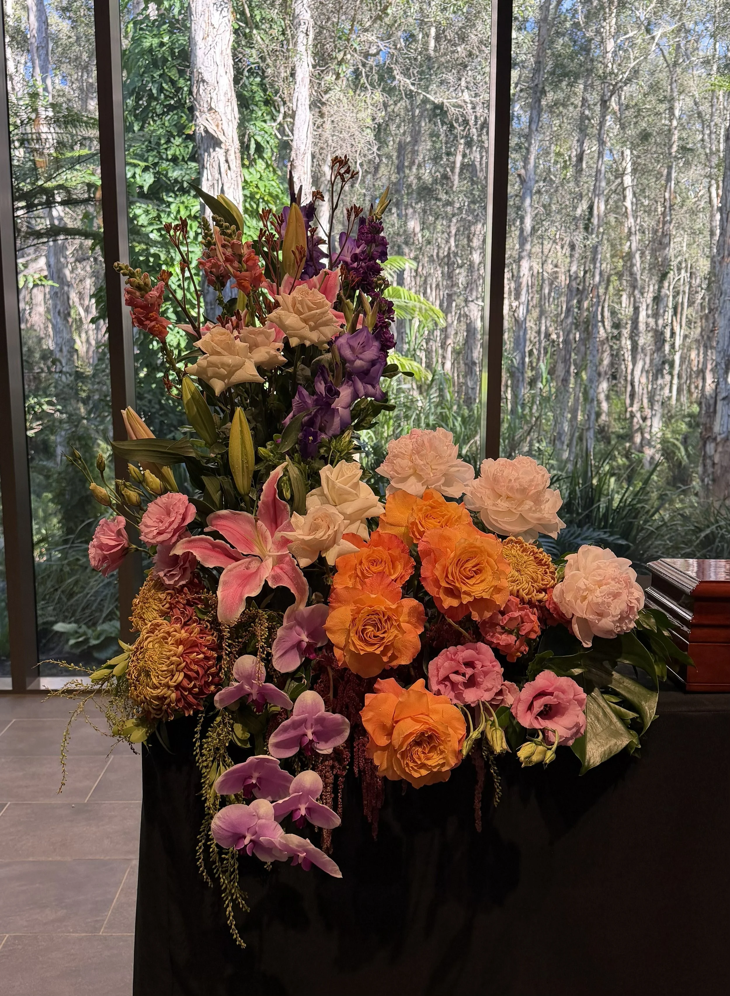 A colorful bouquet of flowers, including orchids, roses, lilies, and other blooms, in front of large windows with a view of a forested area outside.