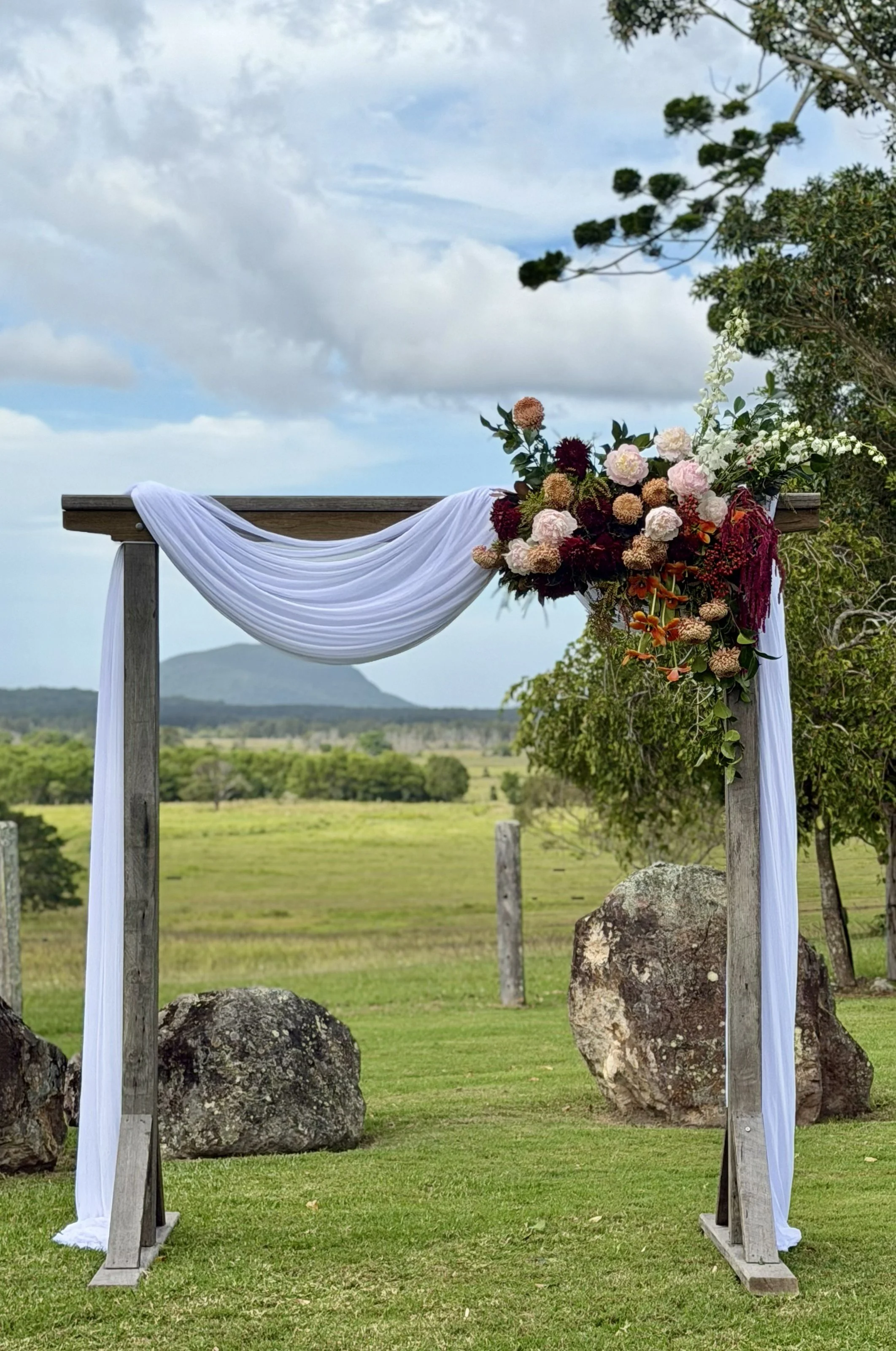 A wooden wedding arch decorated with white fabric and a large floral arrangement on the right side, set outdoors on green grass with trees and a cloudy sky in the background.