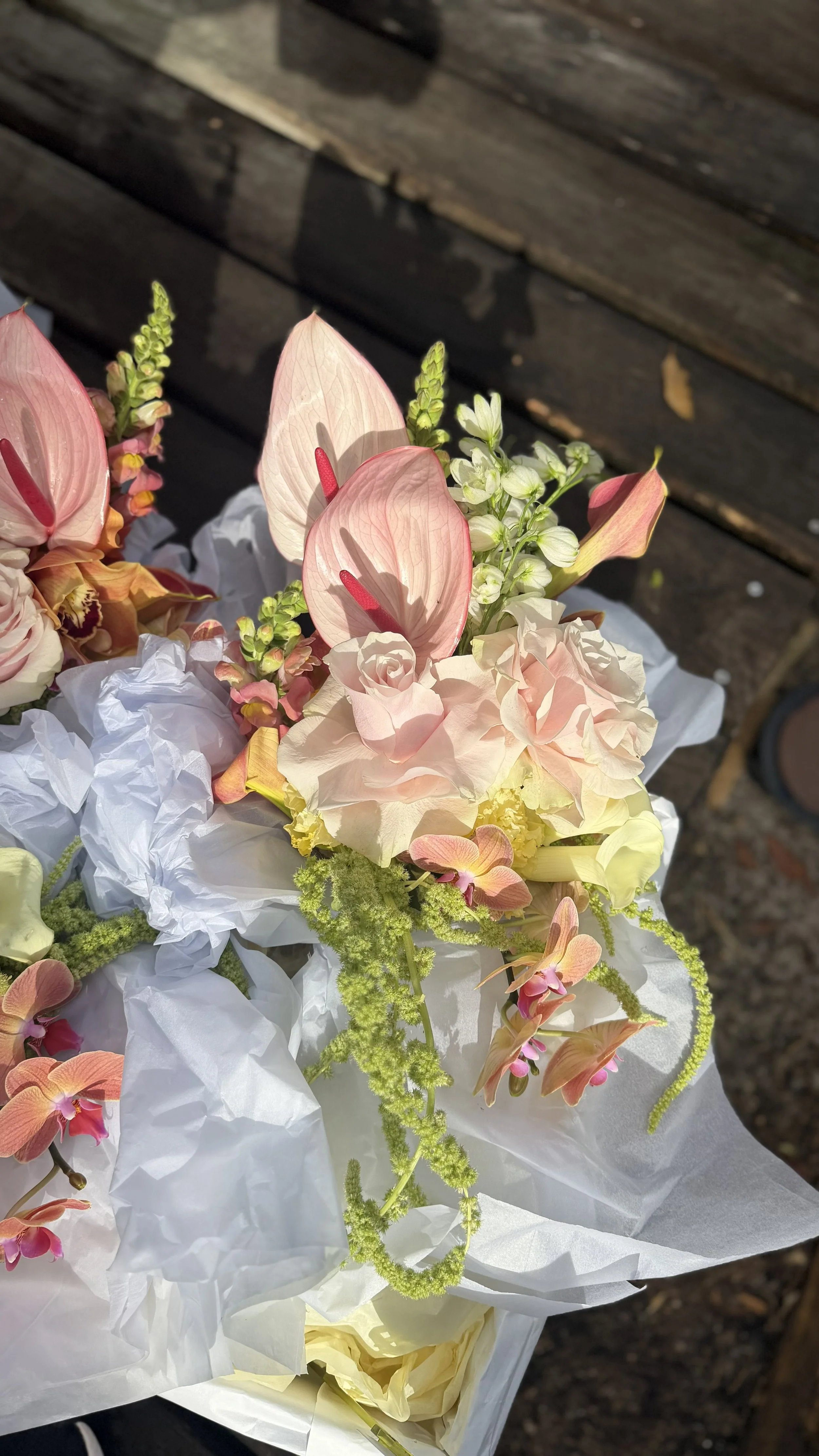 Bouquet of light pink and cream flowers wrapped in white paper, set against a dark wooden background.
