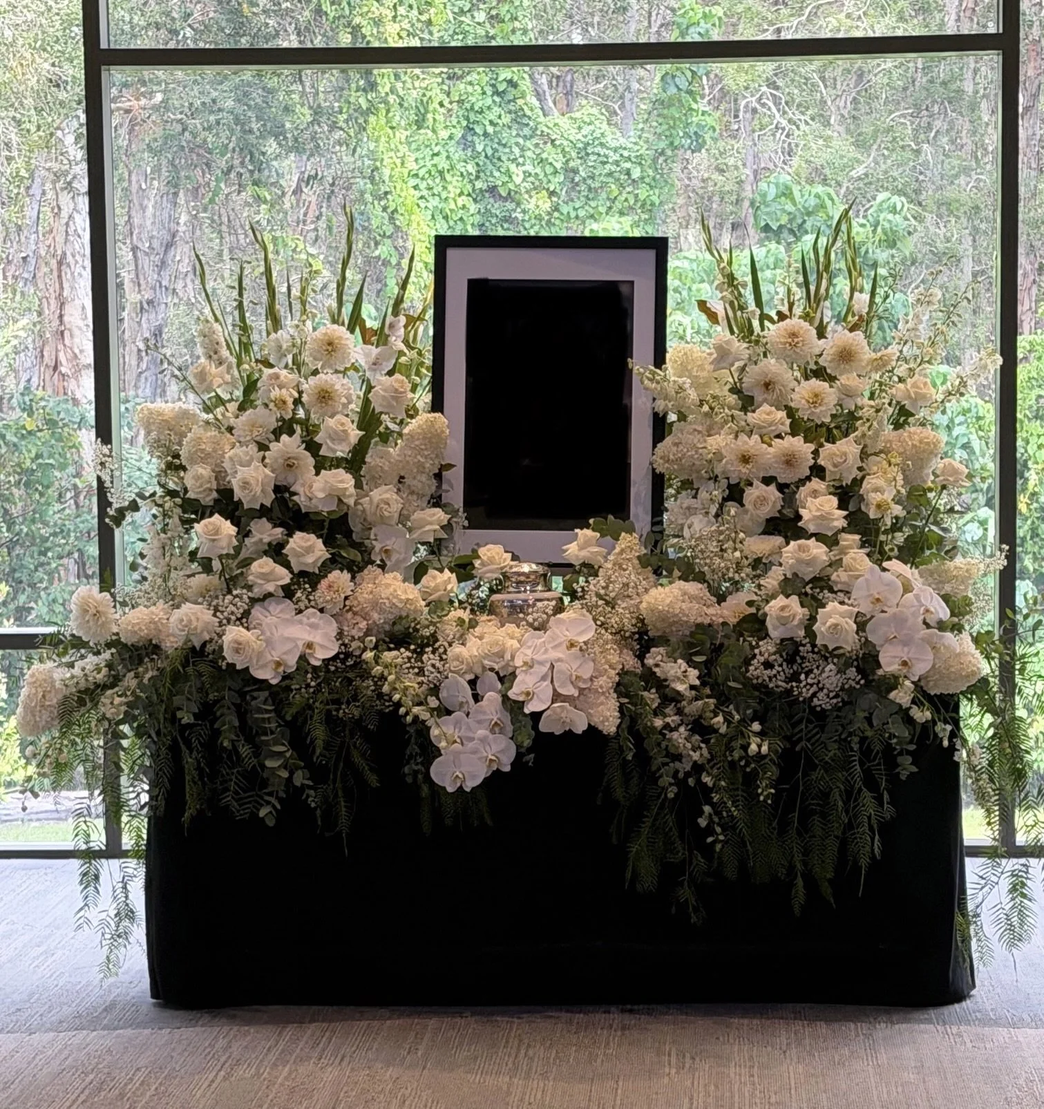 A funeral setup with white and pastel flowers on a black table against a glass wall showing lush green plants outside, with candles and a wooden casket at the center.