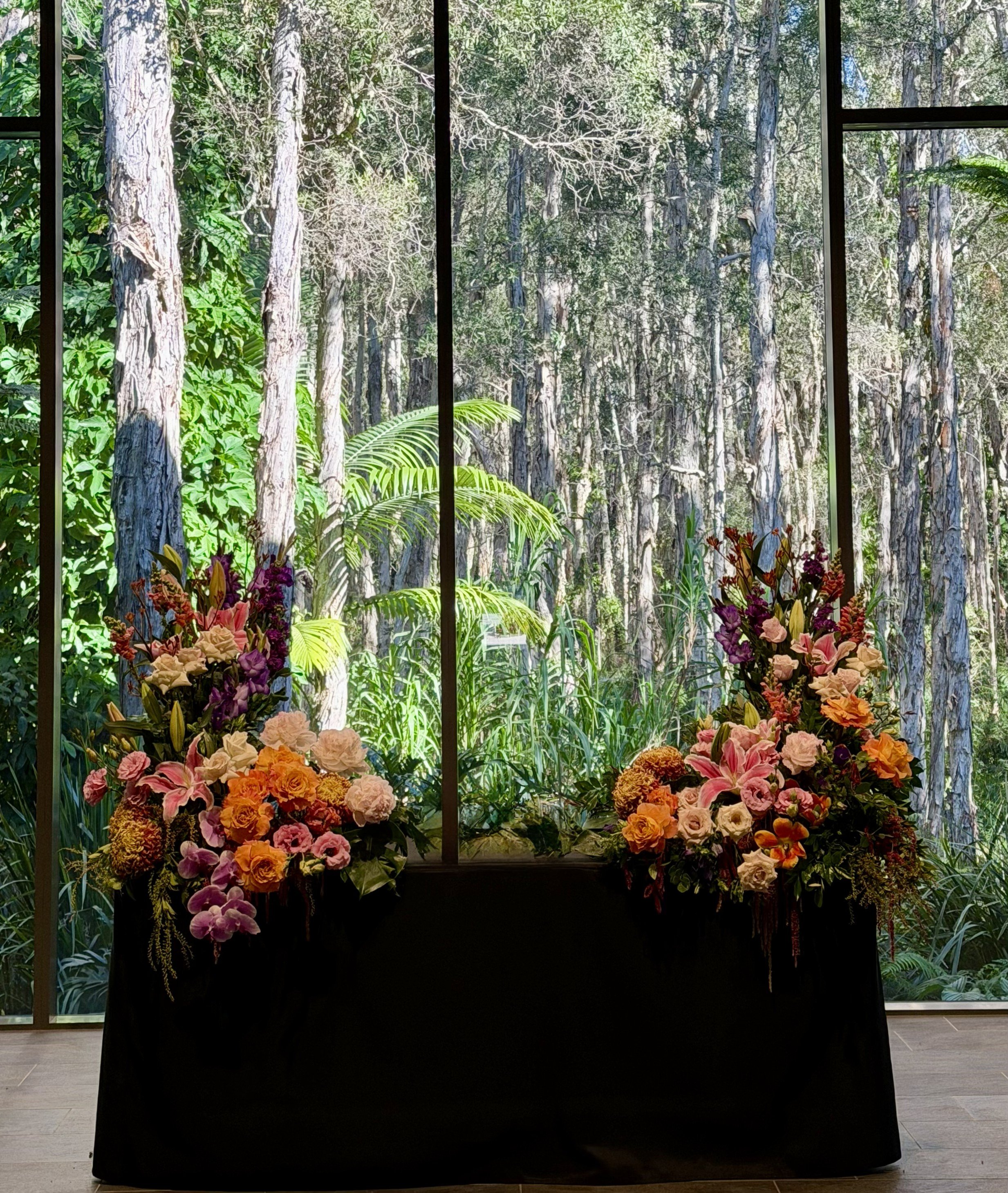 Indoor floral display with a large window showing a forest of tall trees with green foliage outside.
