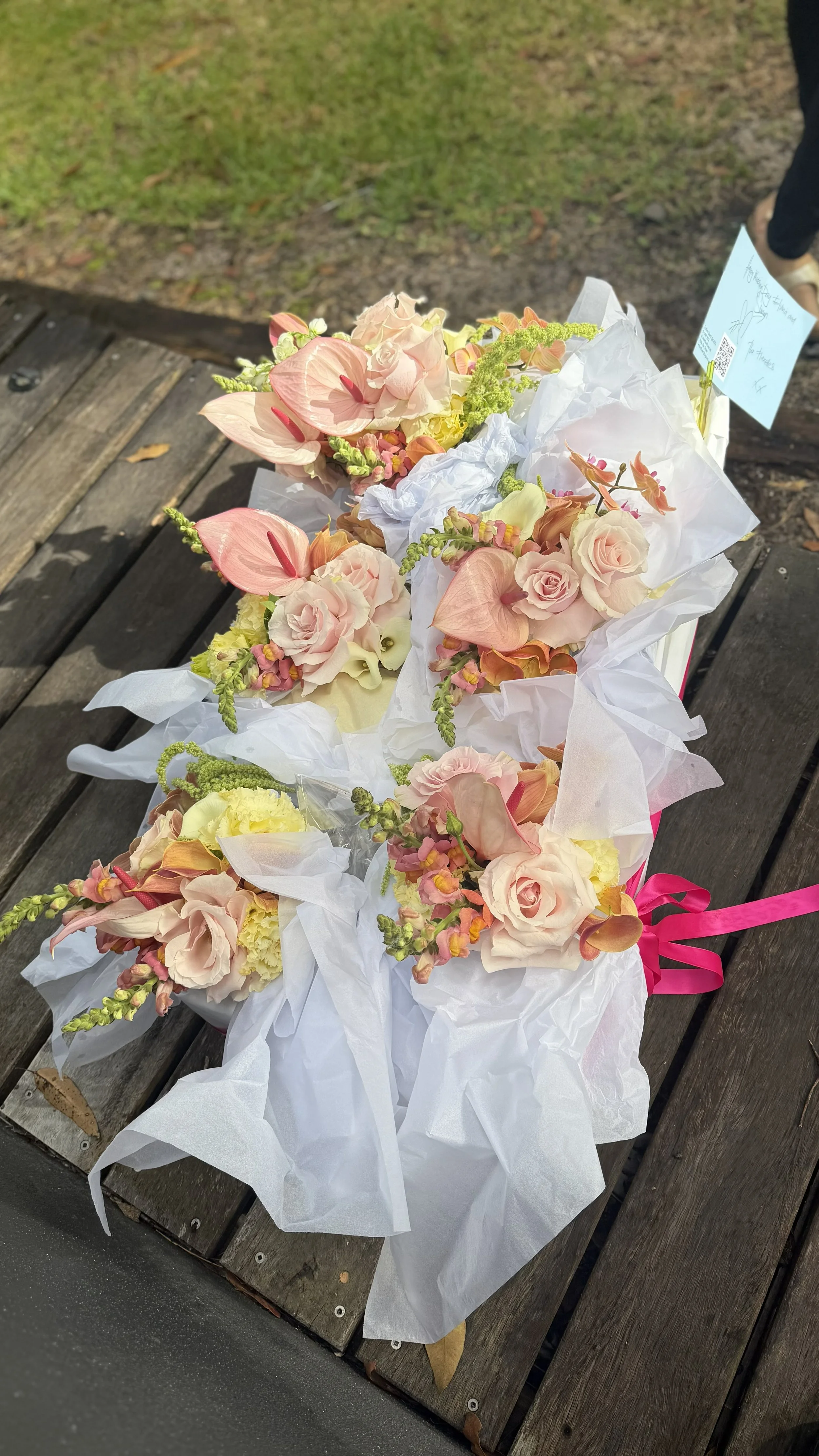 Bouquet of pink, peach, and cream flowers wrapped in white paper with a pink ribbon, placed on a wooden bench outdoors.