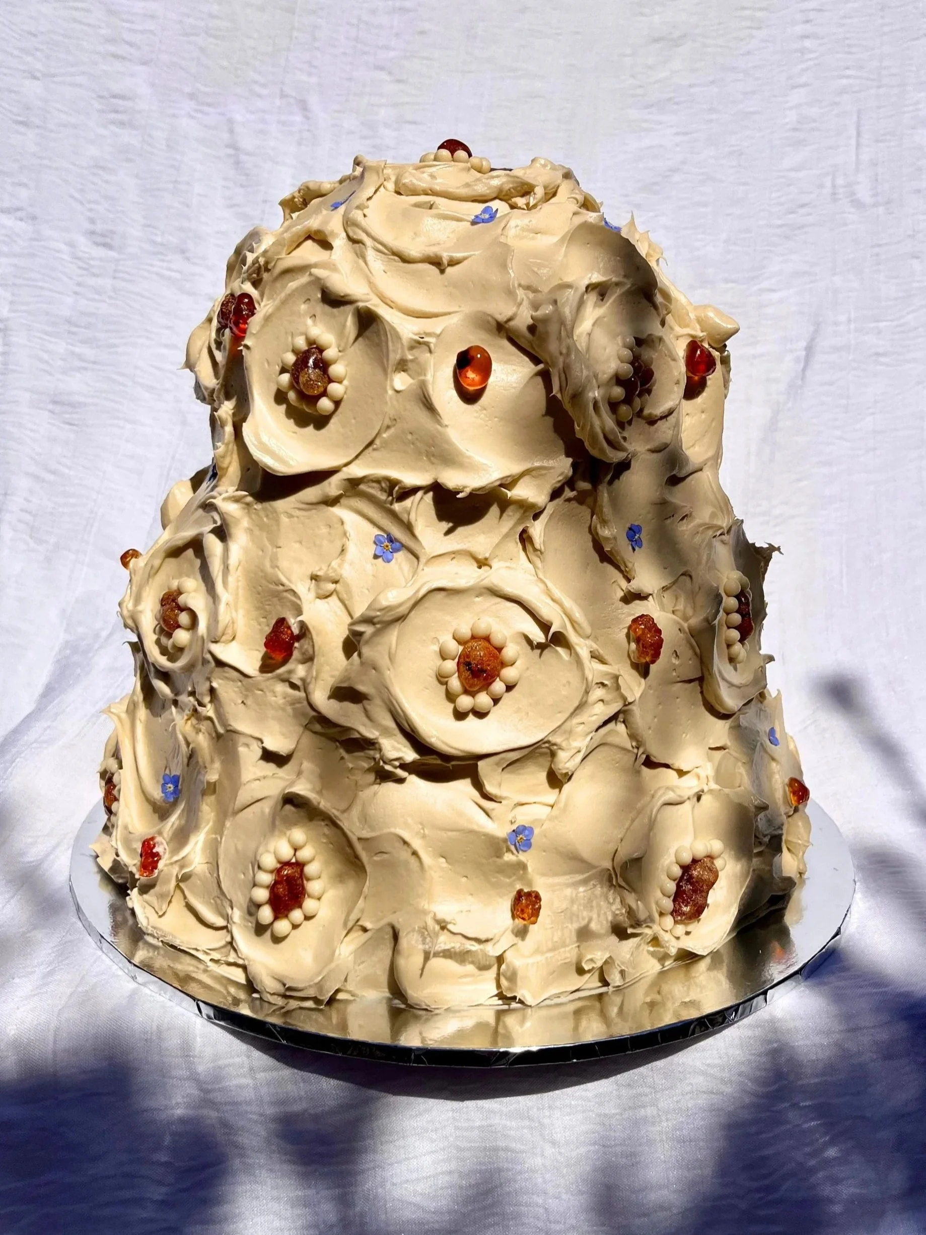 A wedding cake decorated with taupe buttercream, small blue forget-me-nots, amber stones, and white chocolate crispy pearls placed on a reflective silver surface.